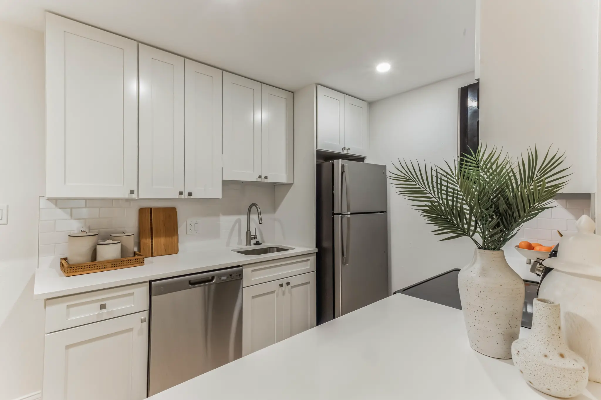 Modern white galley kitchen with shaker cabinets, stainless steel refrigerator and dishwasher, white quartz countertops, subway tile backsplash, recessed lighting, and decorative plant on island counter.