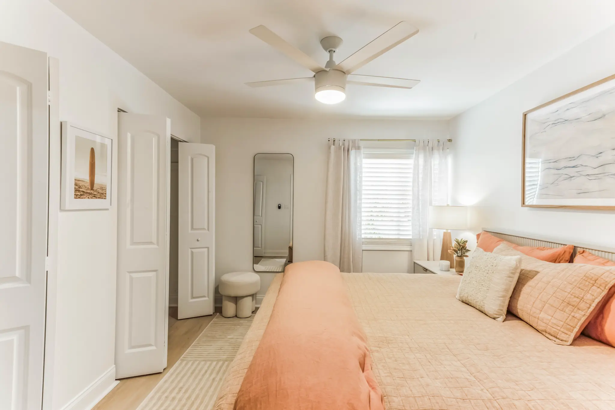 Bright modern bedroom with peach and cream bedding, ceiling fan, large window with sheer curtains, full-length mirror, light wood flooring, and minimalist wall art.