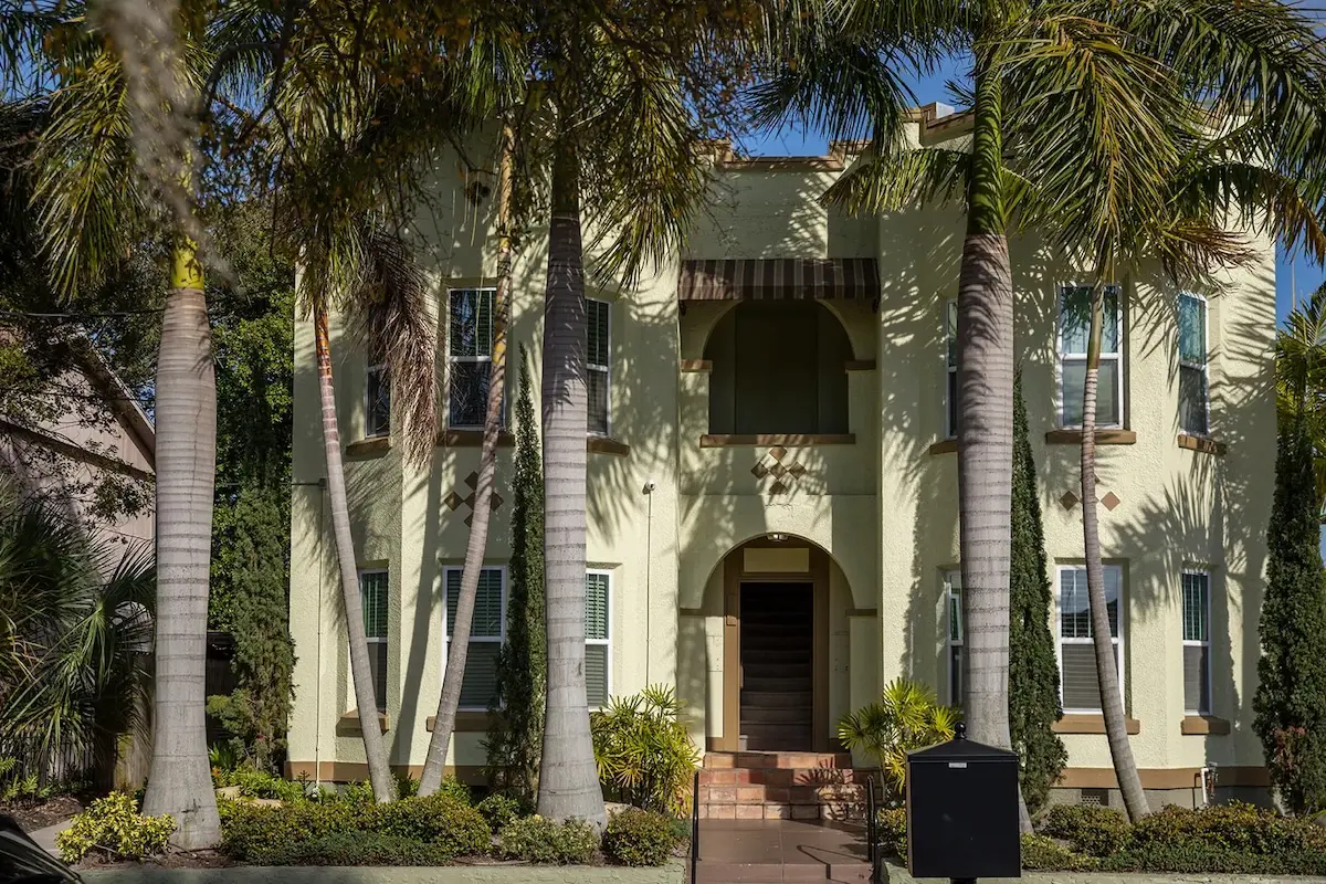 A yellow stucco apartment building surrounded by tall palm trees on a sunny day.