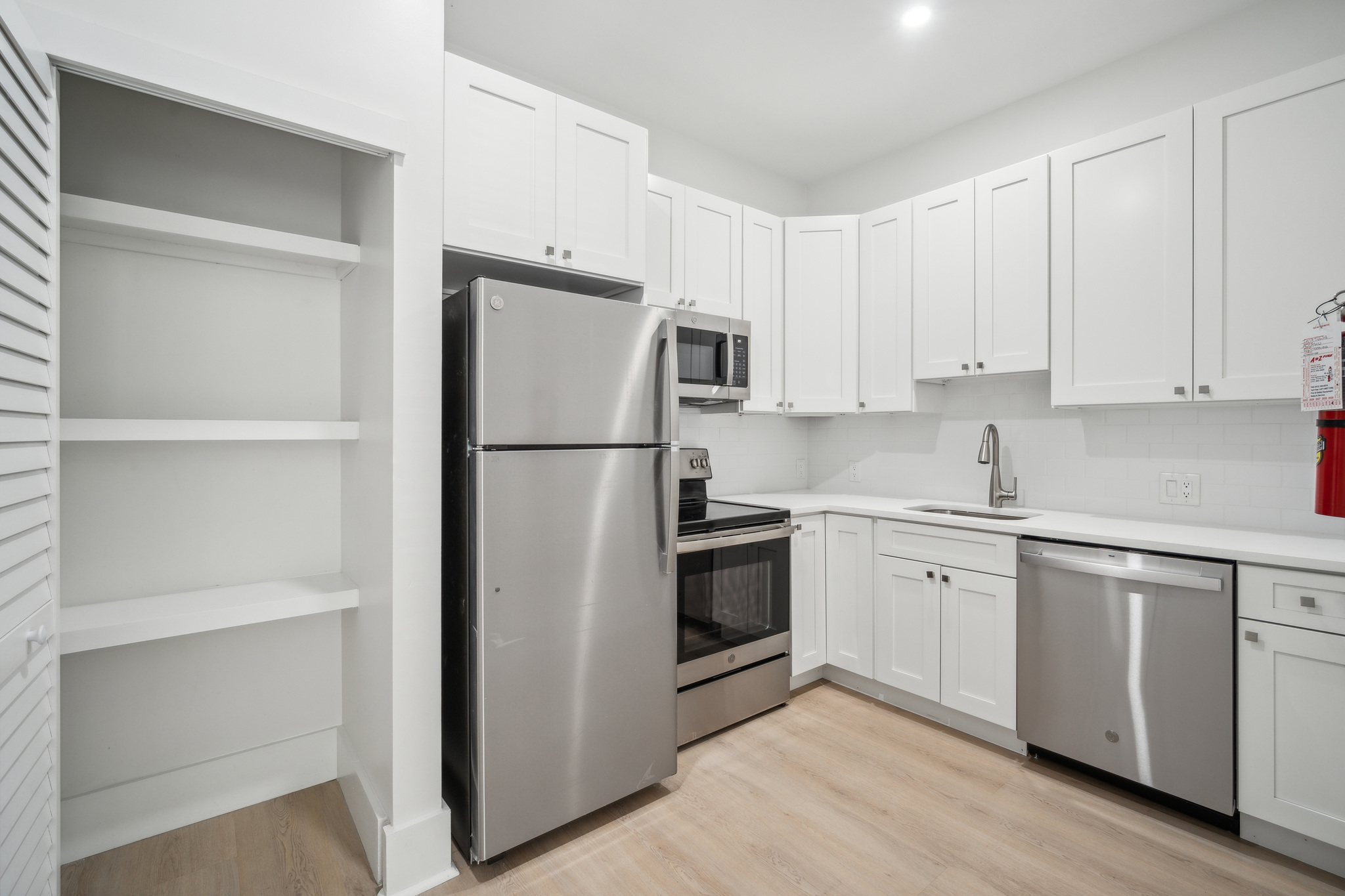 Modern white kitchen with shaker cabinets, stainless steel refrigerator, oven and microwave, sink, dishwasher, light wood floors, and storage.