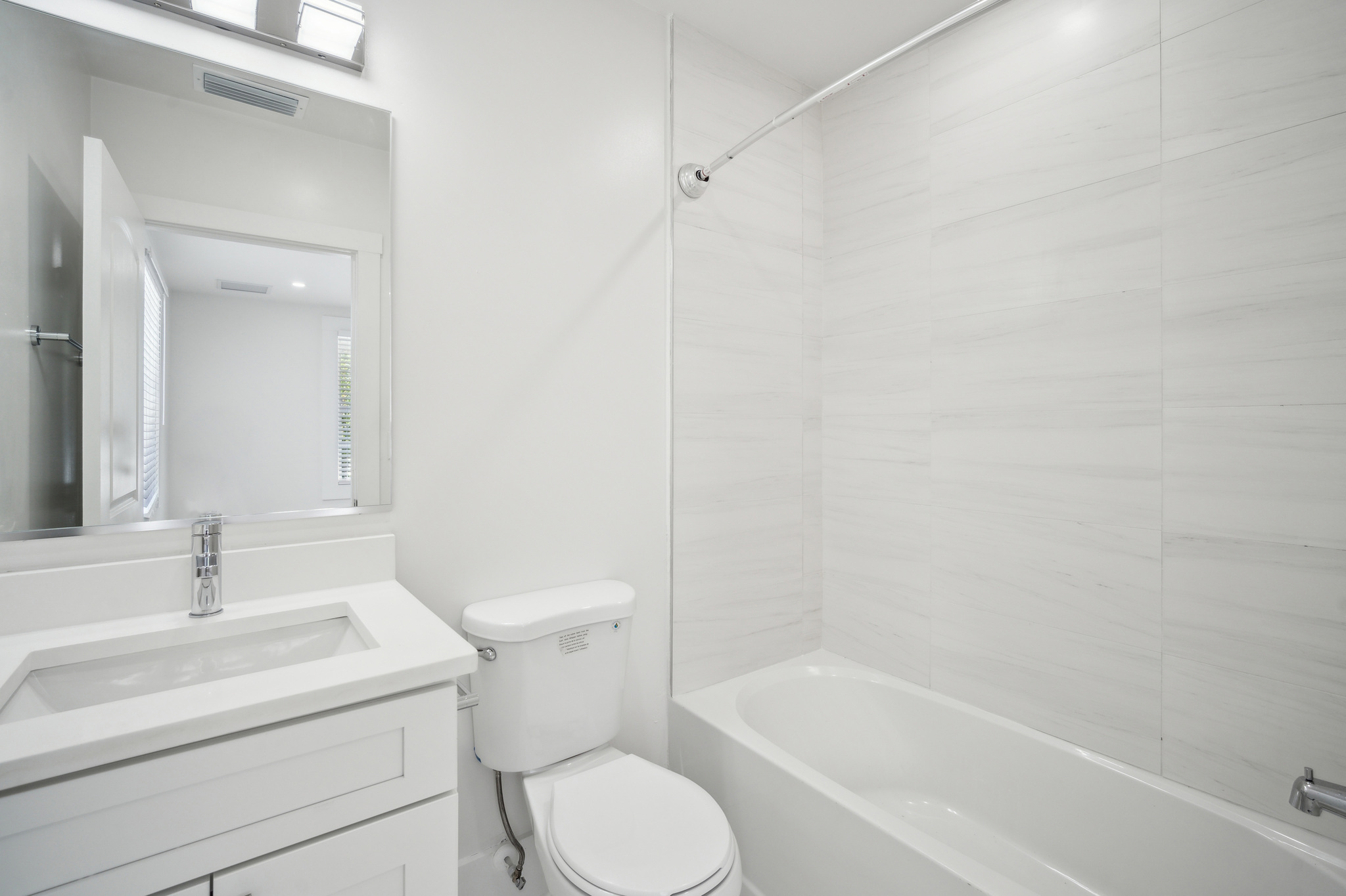 Modern white bathroom with bathtub and shower combo, tiled walls, single-sink vanity with quartz countertop, large mirror, chrome fixtures, and vanity lighting.