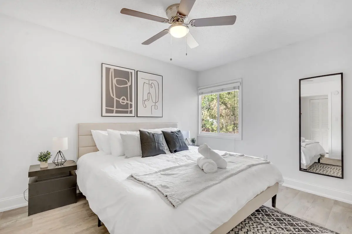 Bright modern bedroom with ceiling fan, large window, mirror, light wood flooring, and minimalist wall art.