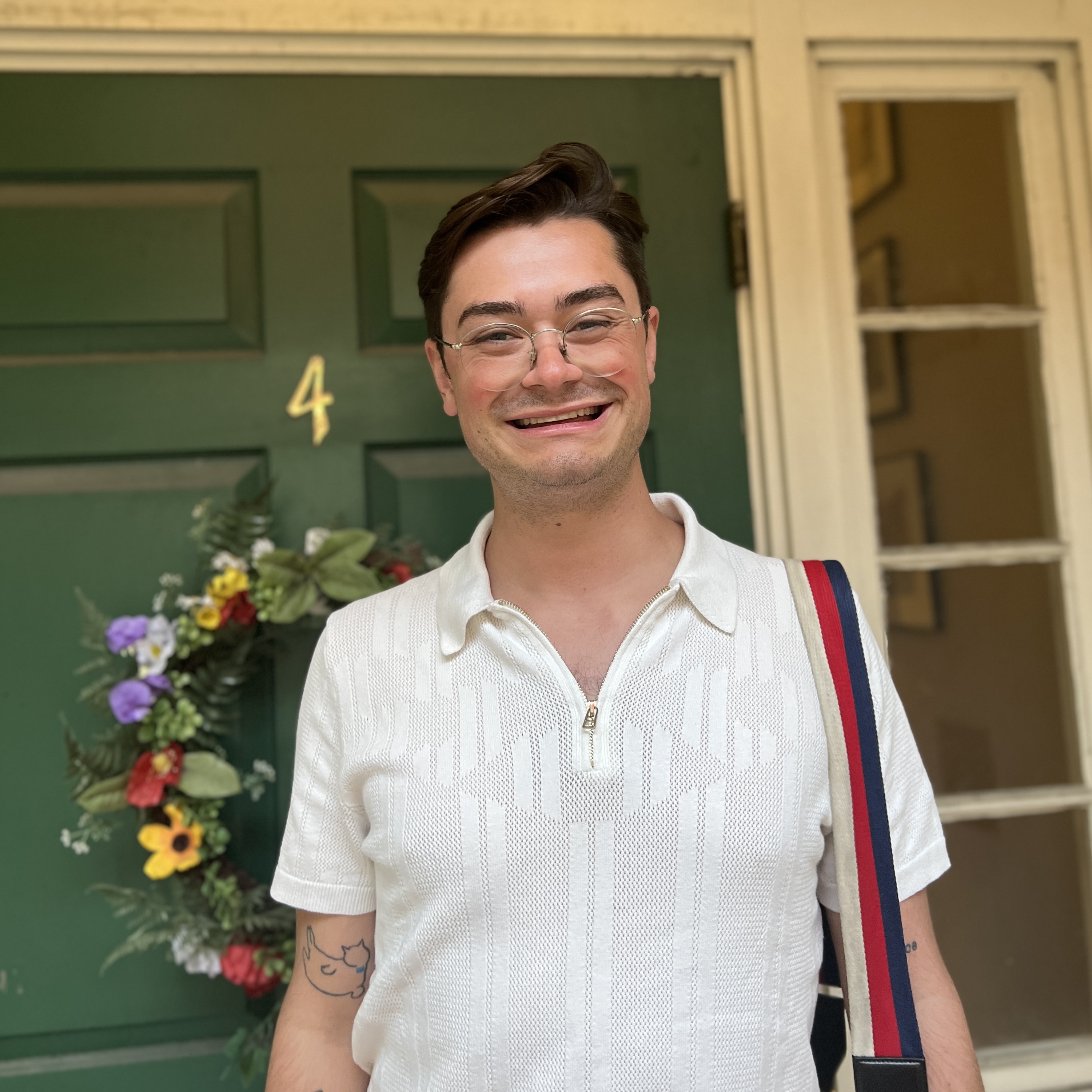 A woman with short brown curly hair and glasses smiling in a art classroom. She is wearing a white top with black floral embroidery.