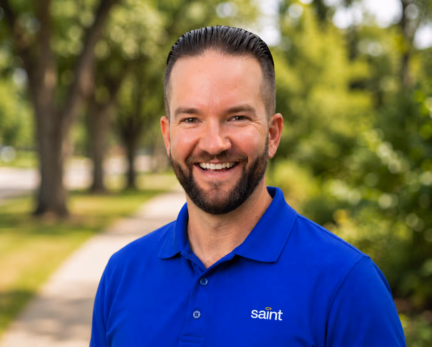 Smiling man with beard wearing a blue polo shirt with 'saint' logo outdoors on a sunny day.