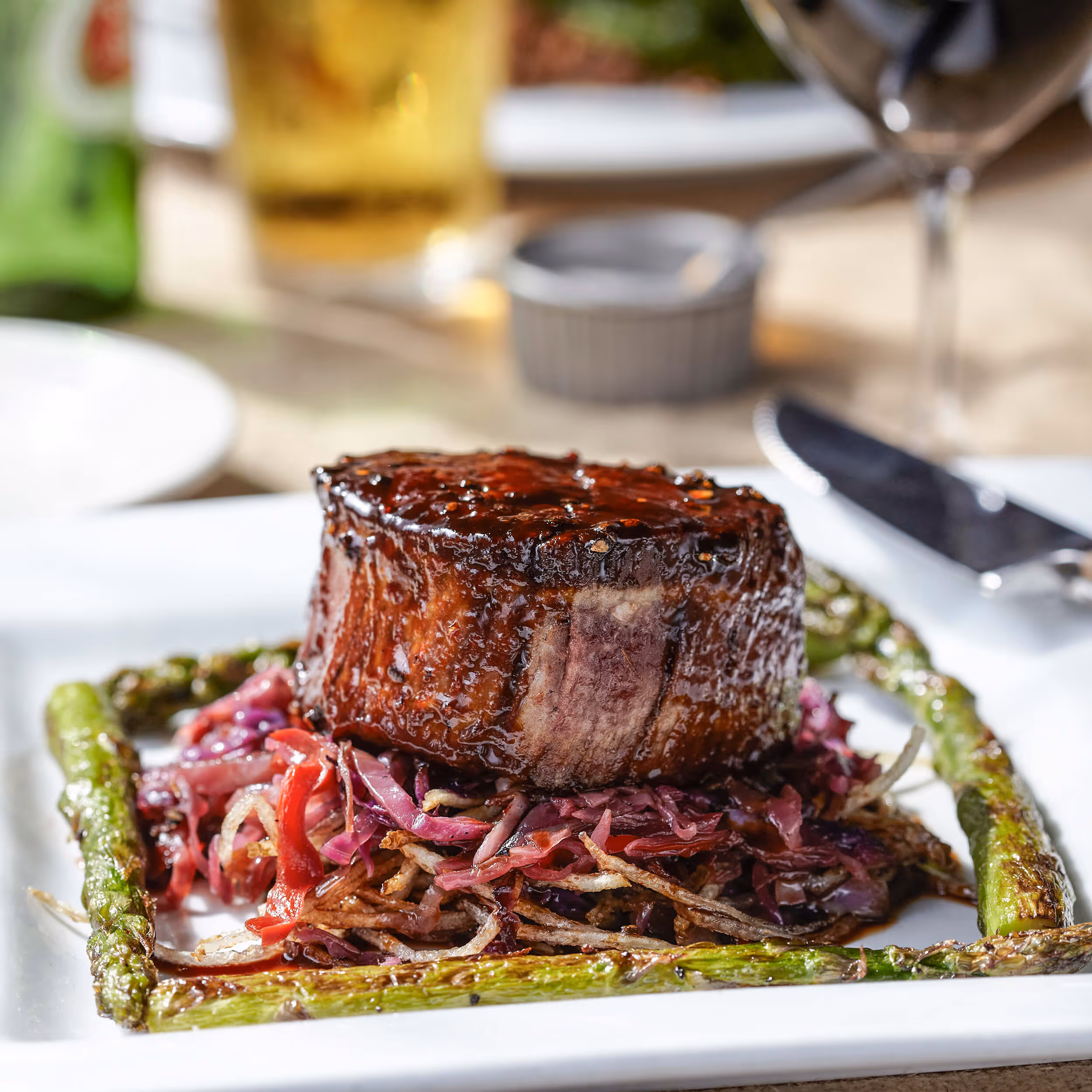 Grilled steak filet wrapped in bacon served on a bed of sautéed shredded vegetables, framed by roasted asparagus on a white plate.