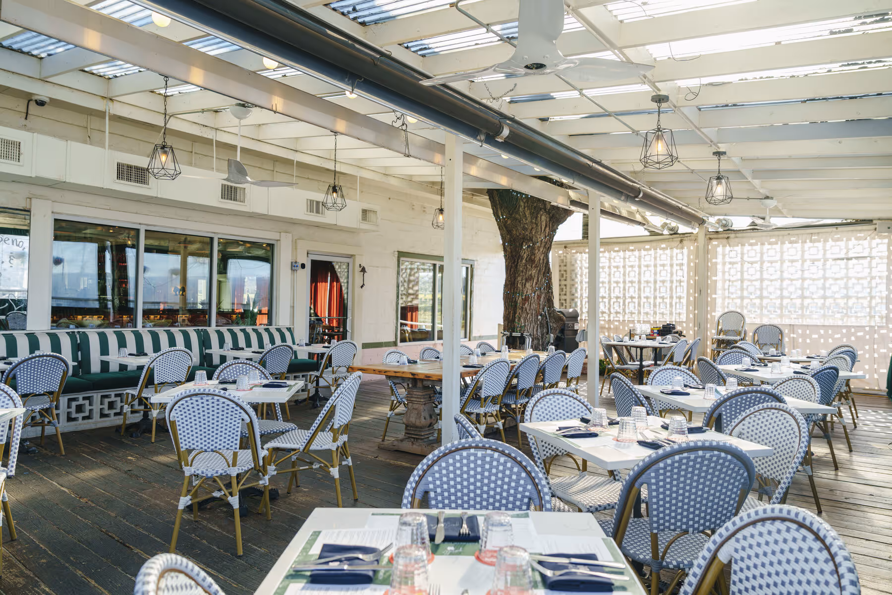 Covered patio dining at Gemelle with blue-and-white bistro chairs, green-striped banquette, and a tree growing through the deck