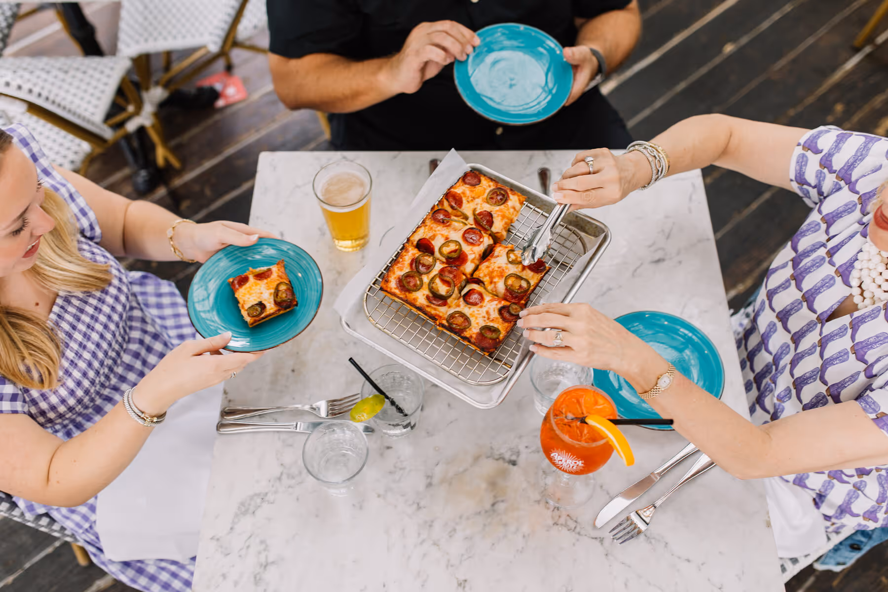 Friends sharing Detroit-style pizza on a marble table with beer, an Aperol Spritz, and aqua plates from above