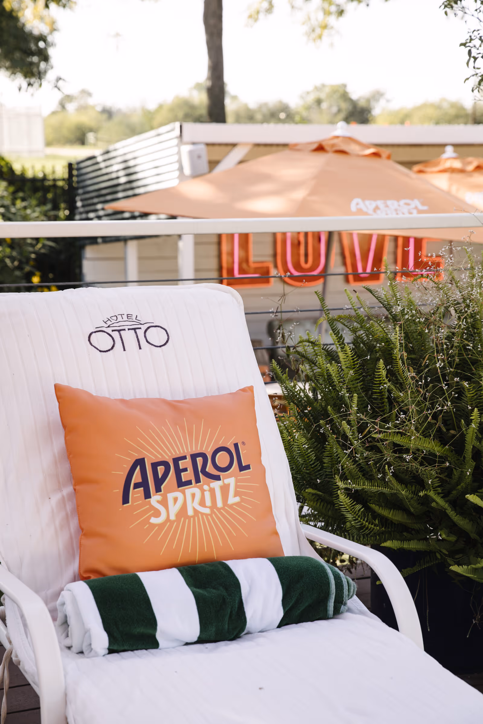 Aperol Spritz throw pillow on a Hotel Otto chaise with a green-striped towel, framed by an Aperol umbrella and the LOVE neon sign