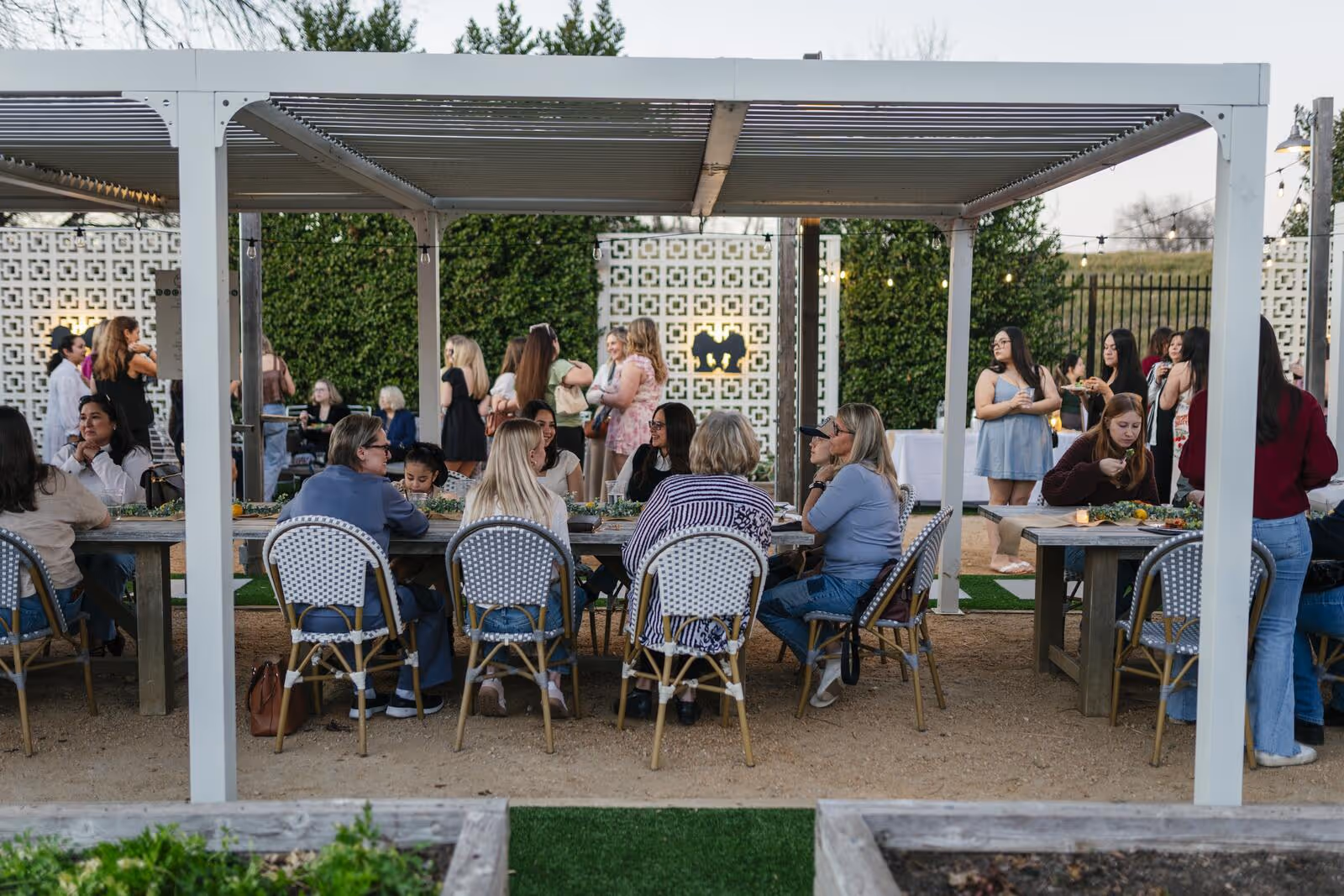 Guests dining at long communal tables under a pergola at dusk, with the Gemelle silhouette panel in the background