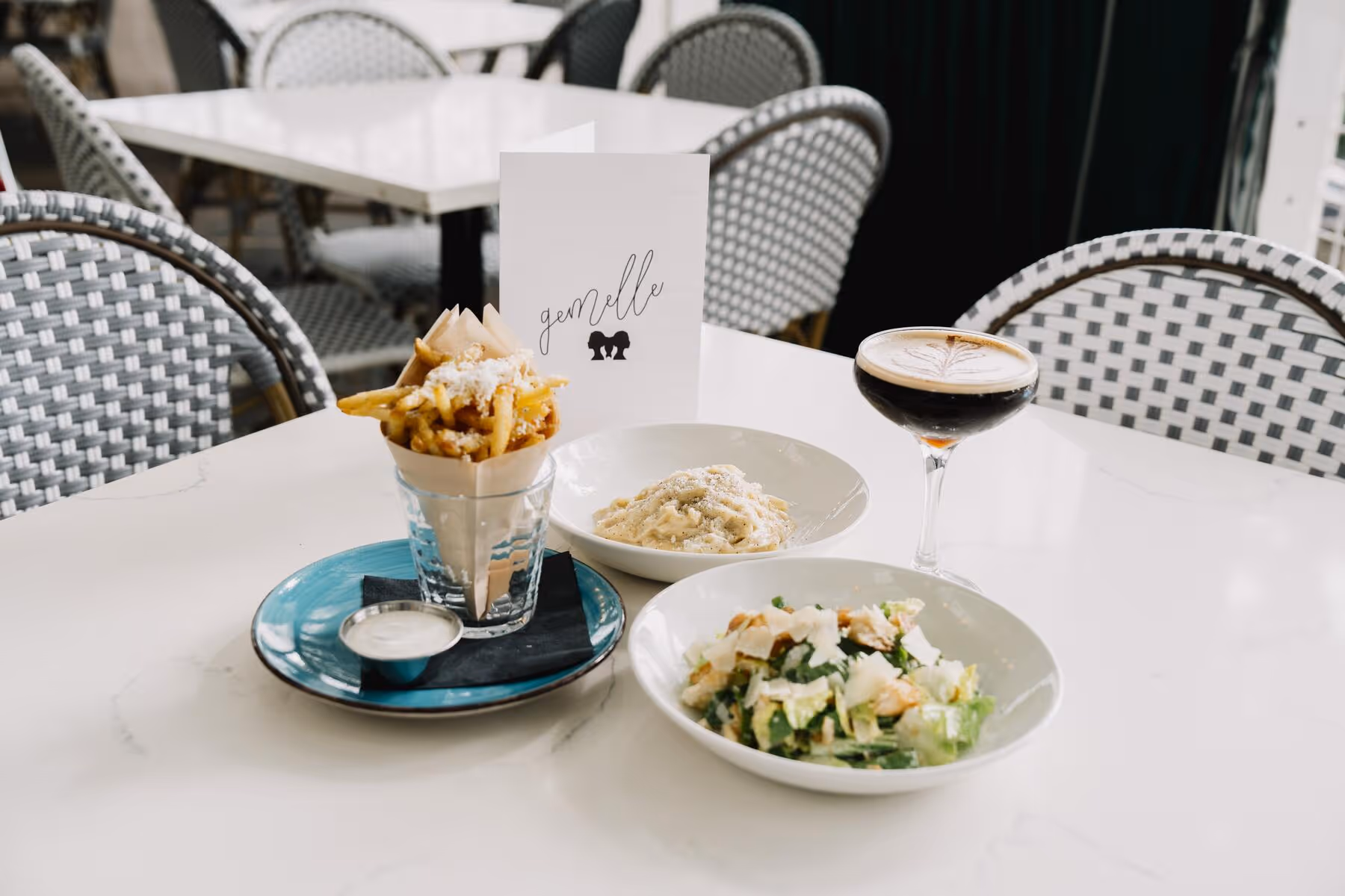 Gemelle table spread with a cone of fries, fettuccine alfredo, Caesar salad, and an espresso martini between blue-and-white bistro chairs