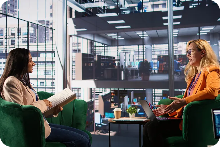 Two professional women sitting in green armchairs facing each other in a modern office with large windows, one holding a book and the other gesturing while using a laptop on a small table between them.