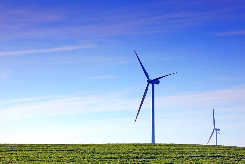 Two wind turbines standing on a green grassy hill beneath a blue sky with soft clouds.