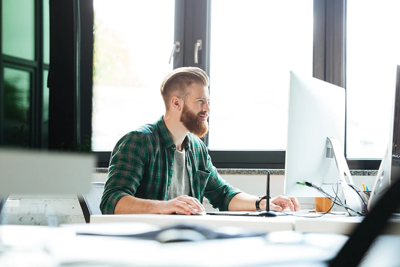 Bearded man in a plaid shirt working on a desktop computer in a bright office.