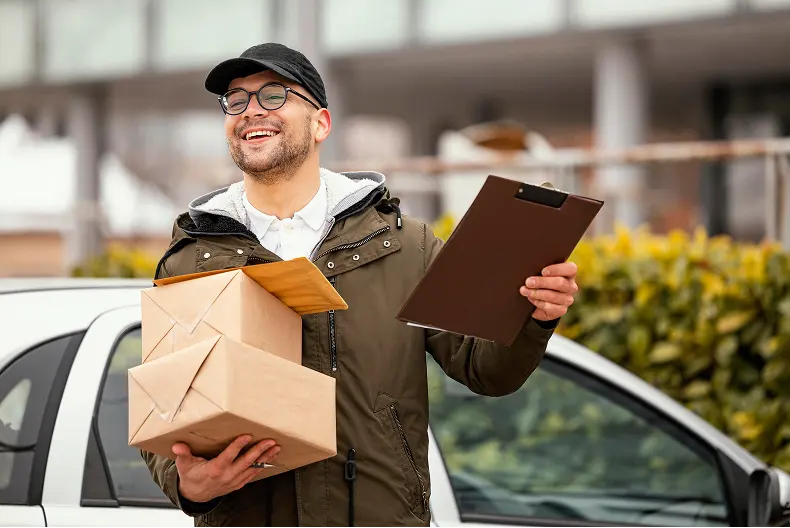 Smiling delivery man holding three cardboard boxes and a clipboard standing next to a white car outdoors.
