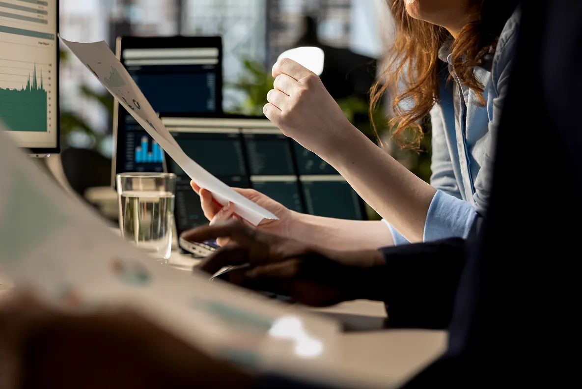 Close-up of two people working at a desk with documents and multiple computer screens displaying charts and graphs.