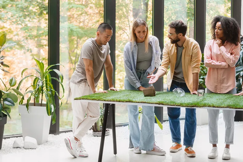 Four diverse young adults standing around a table with artificial grass, discussing eco-friendly products in a bright room with large windows and plants.
