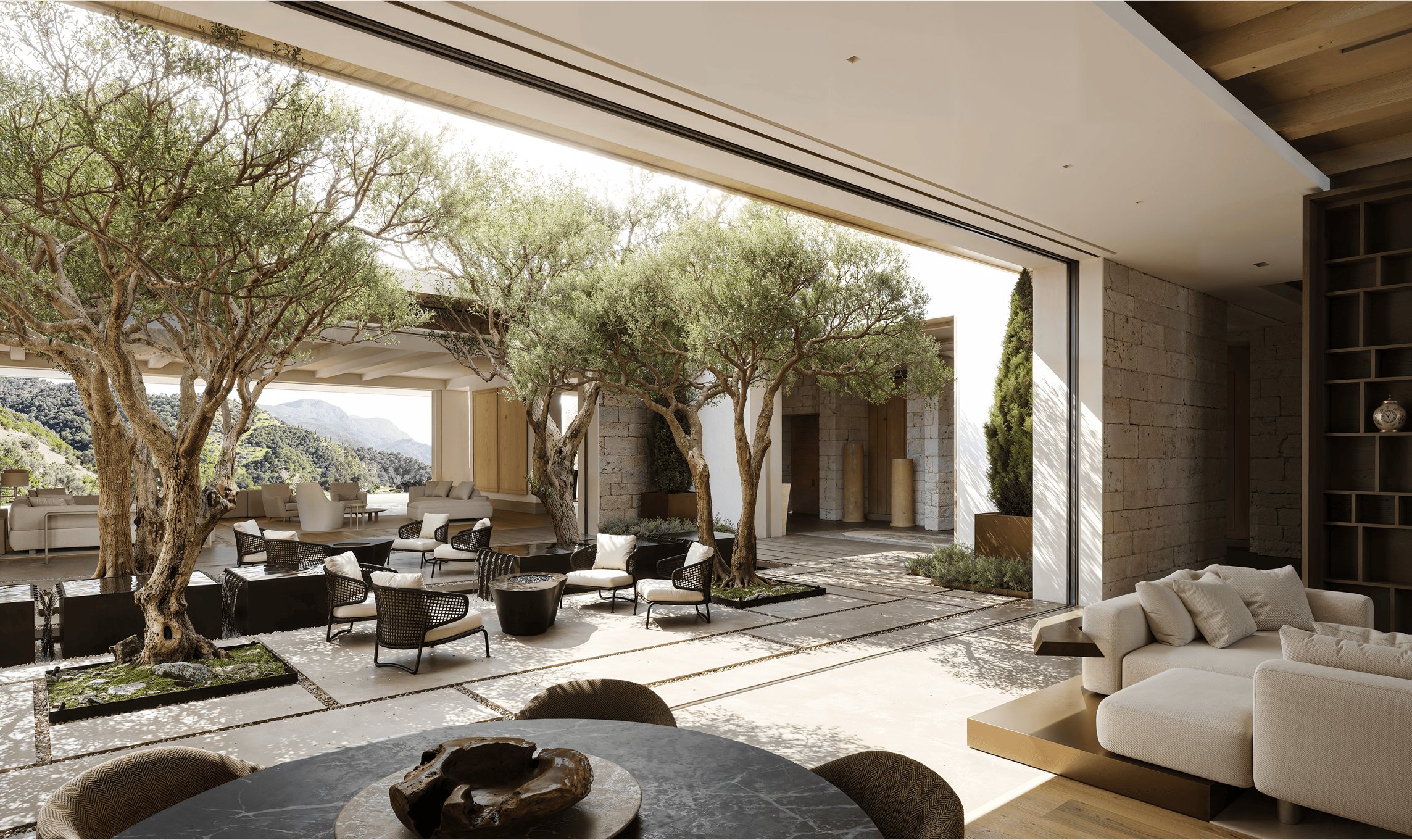 Spacious sunlit modern living area with open walls to an outdoor patio featuring olive trees, black wicker chairs with cushions, and scenic mountain views.