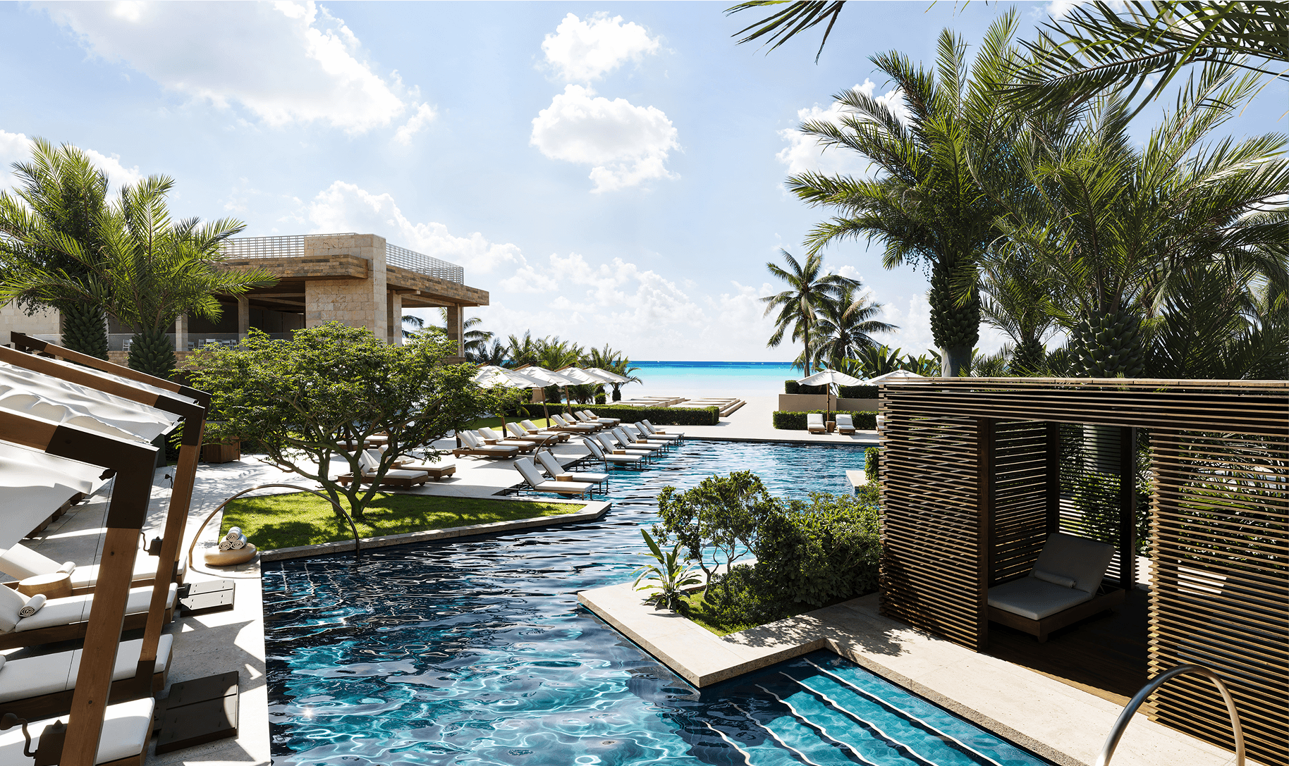Luxury outdoor pool area with lounge chairs, cabanas, palm trees, and a view of the beach and ocean under a sunny sky.