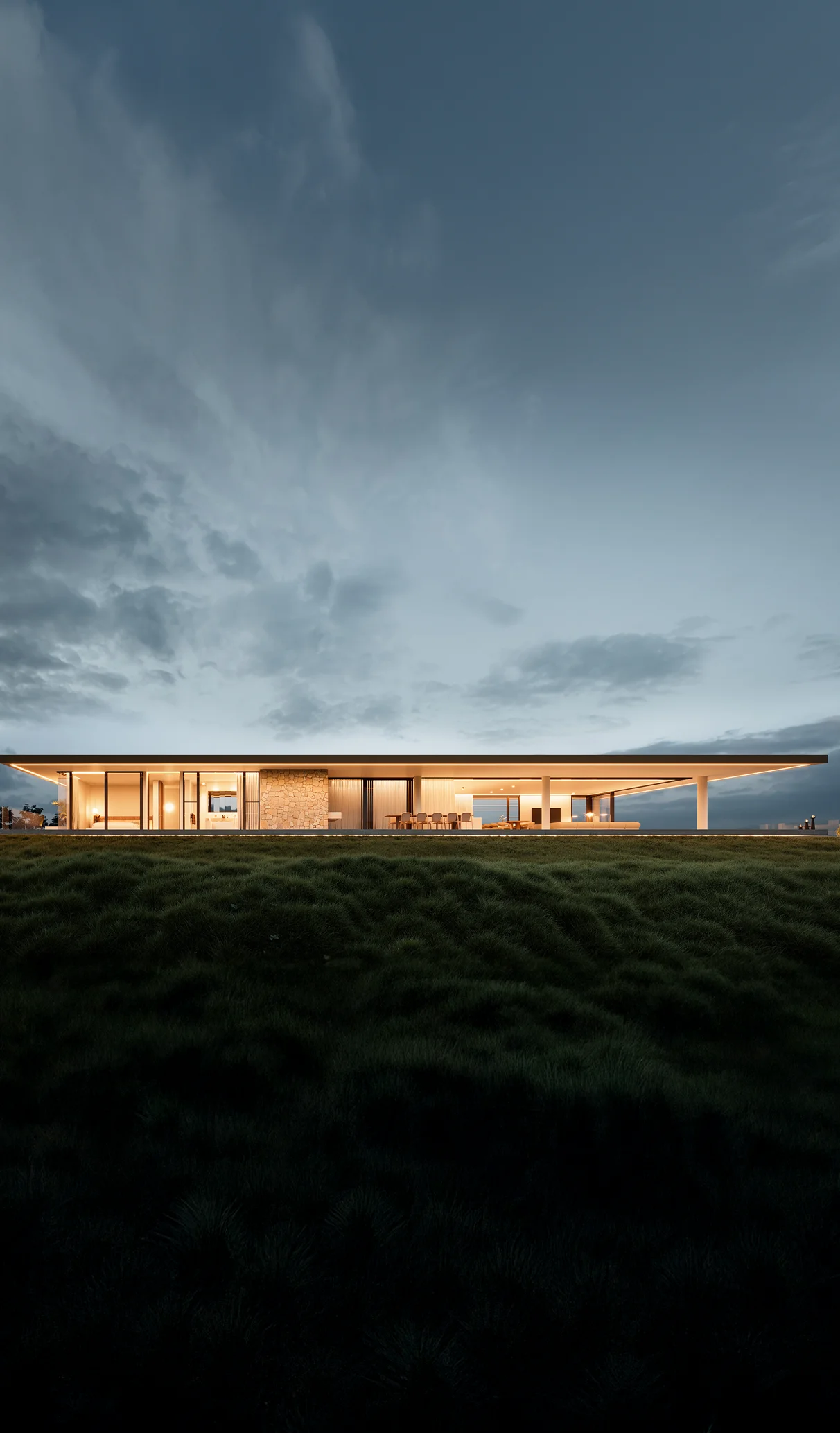 Modern minimalist house with a flat roof and large windows illuminated at dusk, sitting atop a grassy hill under a cloudy sky.