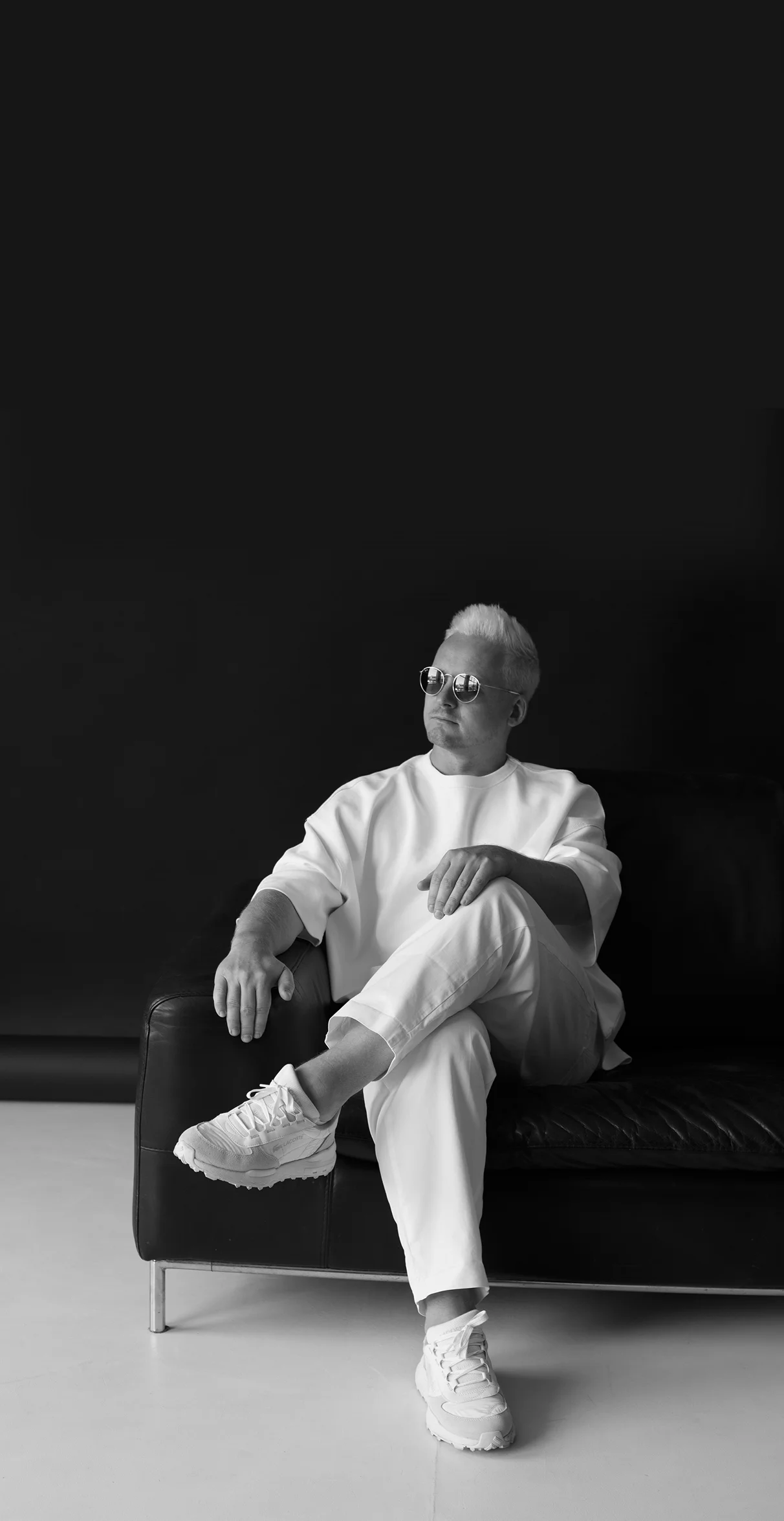 Man with short light hair wearing sunglasses and white clothes sitting with legs crossed on a black leather sofa against a dark background.