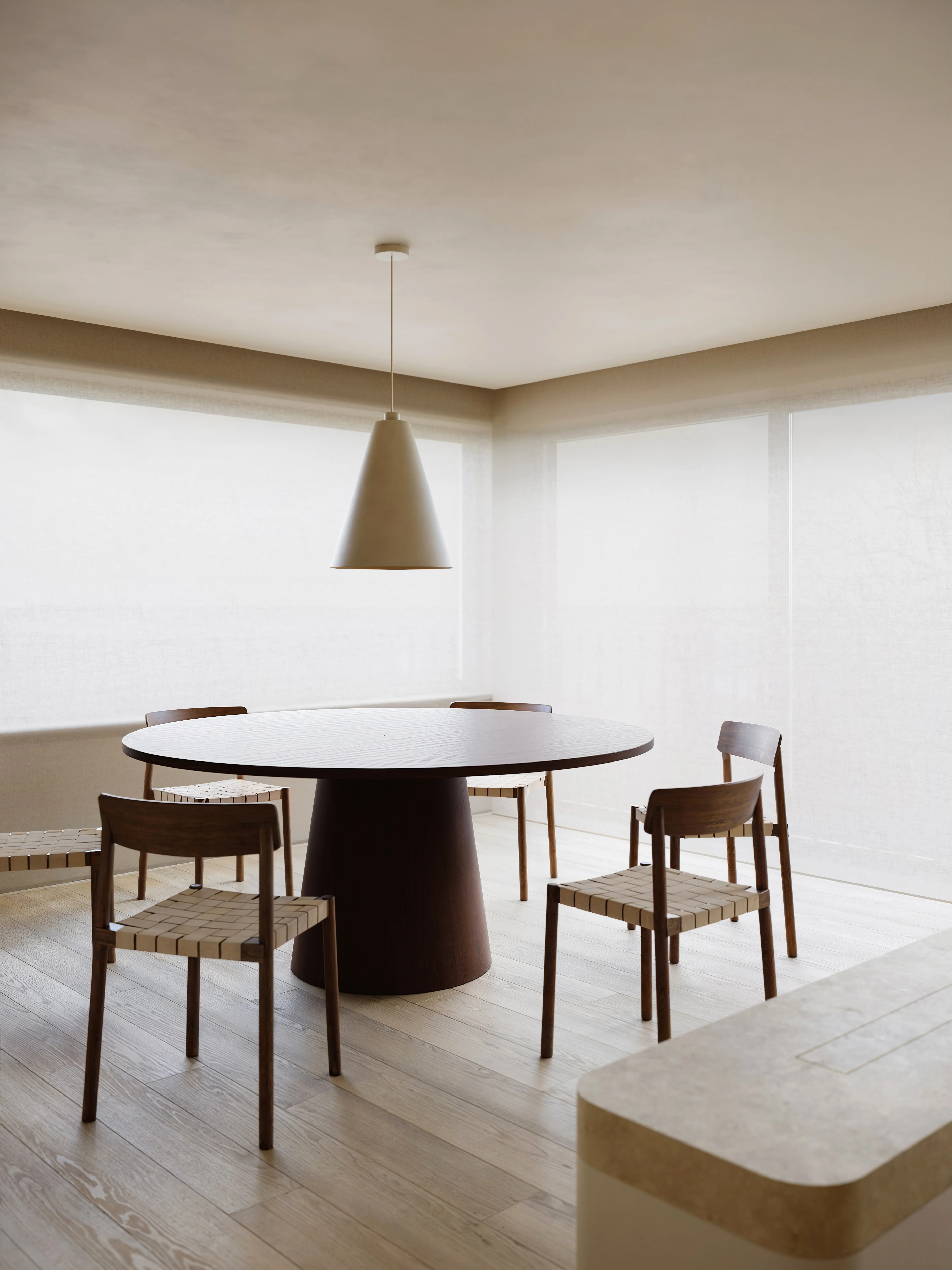 Minimalist dining area with a round dark wooden table, four wooden chairs with woven seats, and large windows covered with sheer blinds.
