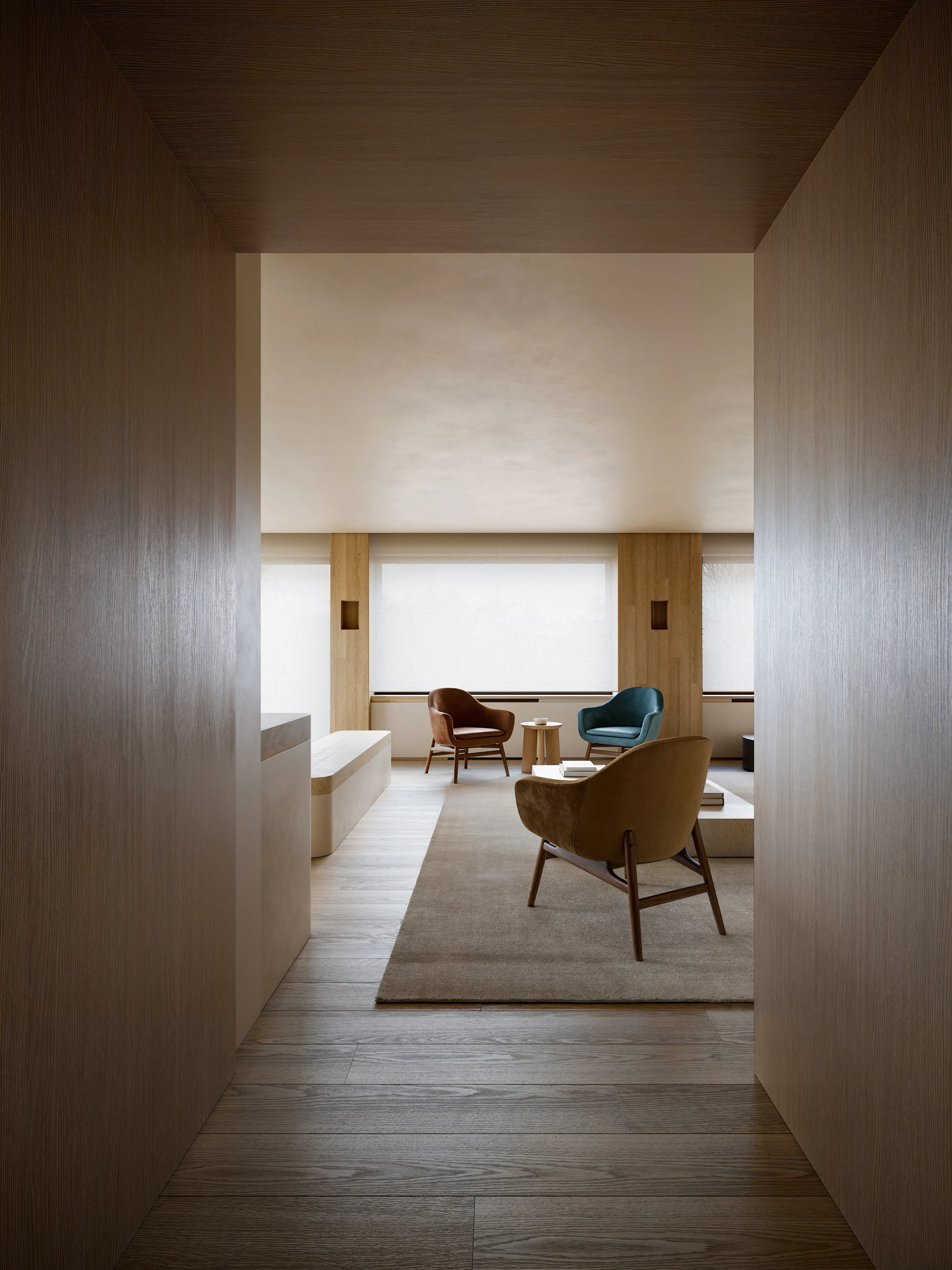 Minimalist living room with brown, teal, and beige armchairs, wooden flooring, and large windows with white blinds.