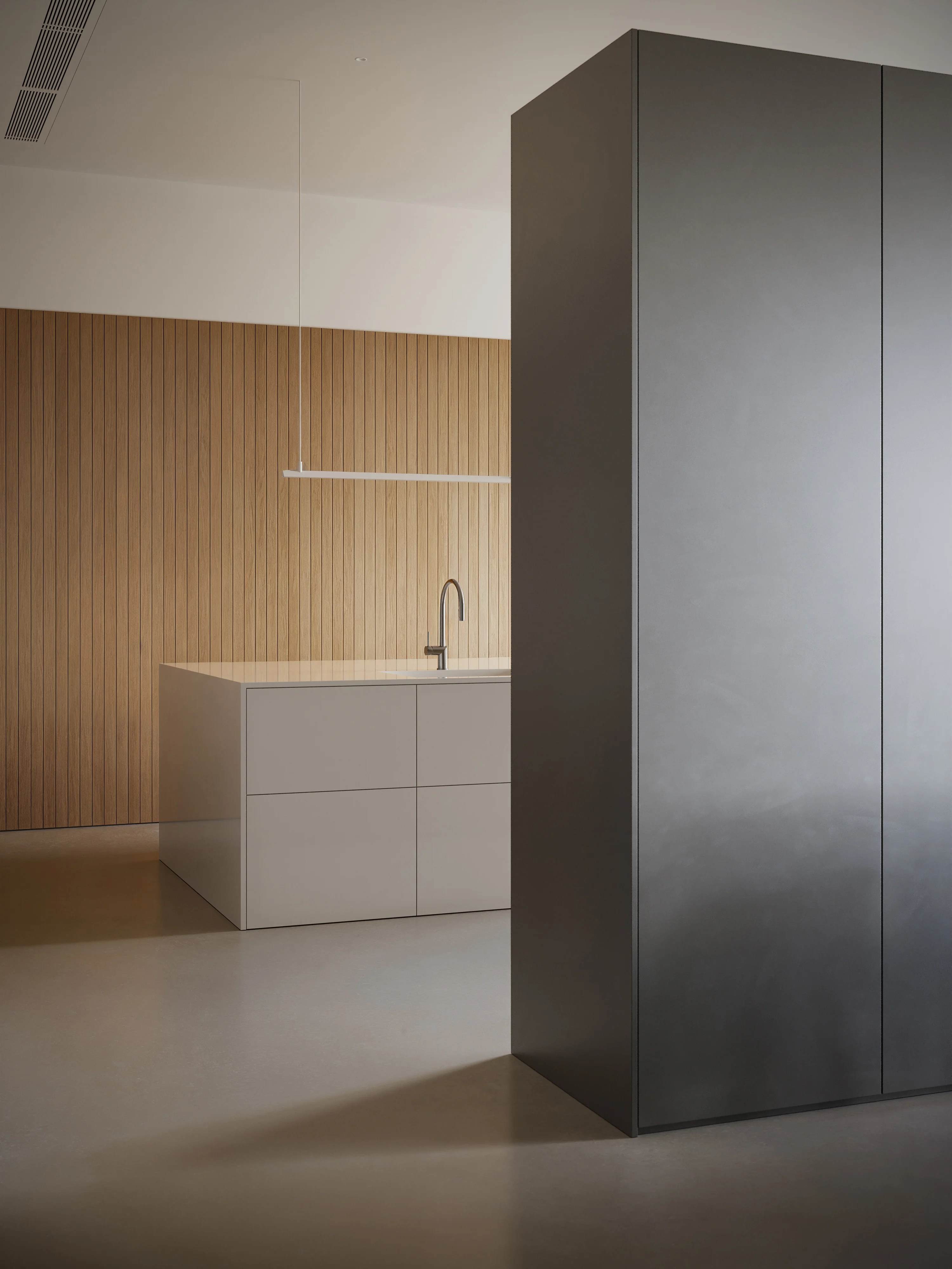 Minimalist kitchen with a large gray cabinet in the foreground, white island with sink and faucet, and vertical wooden panel wall in the background.