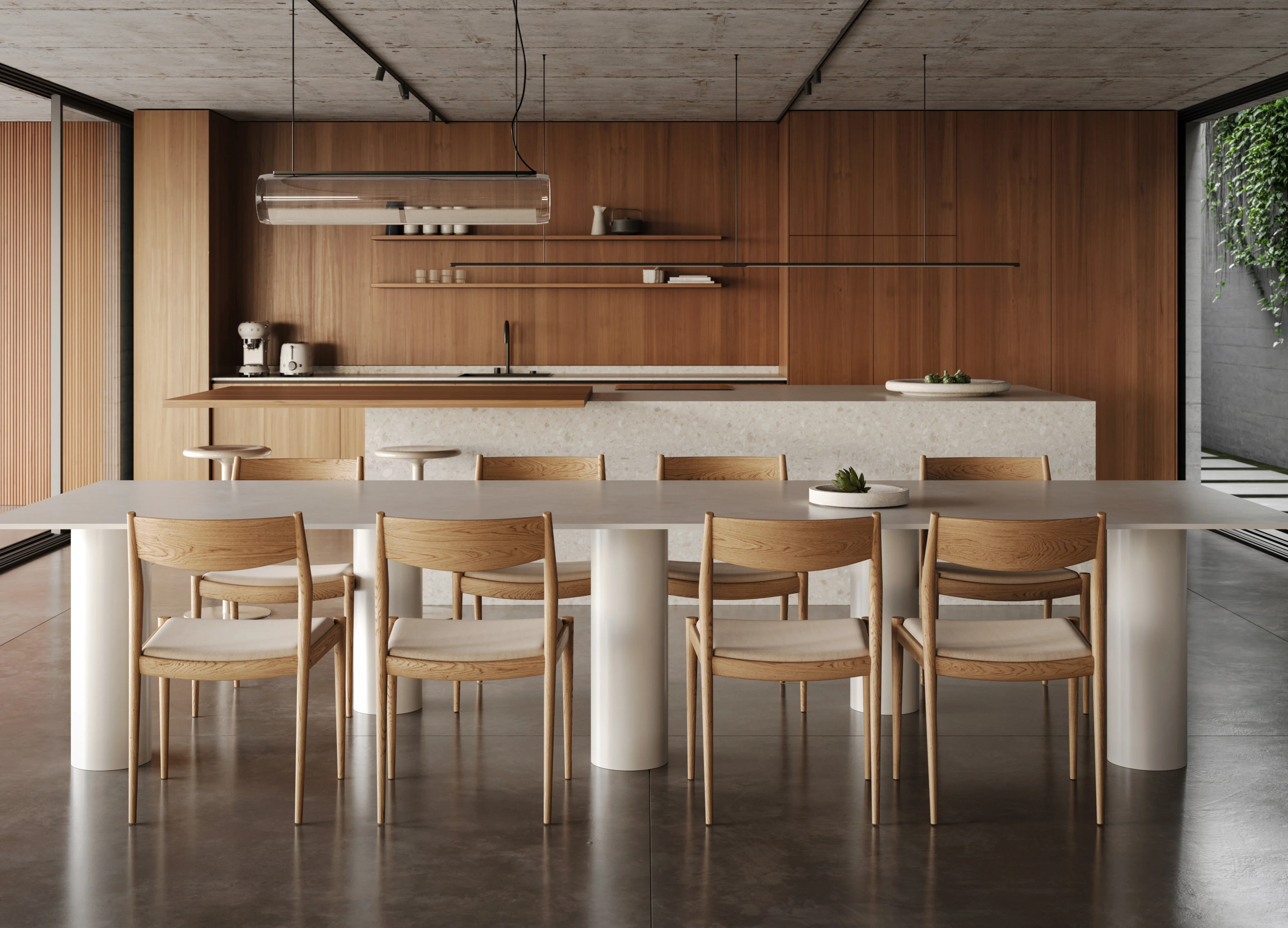Modern dining area with a large white table supported by cylindrical legs and wooden chairs, set against a kitchen with wooden cabinetry and minimalist decor.