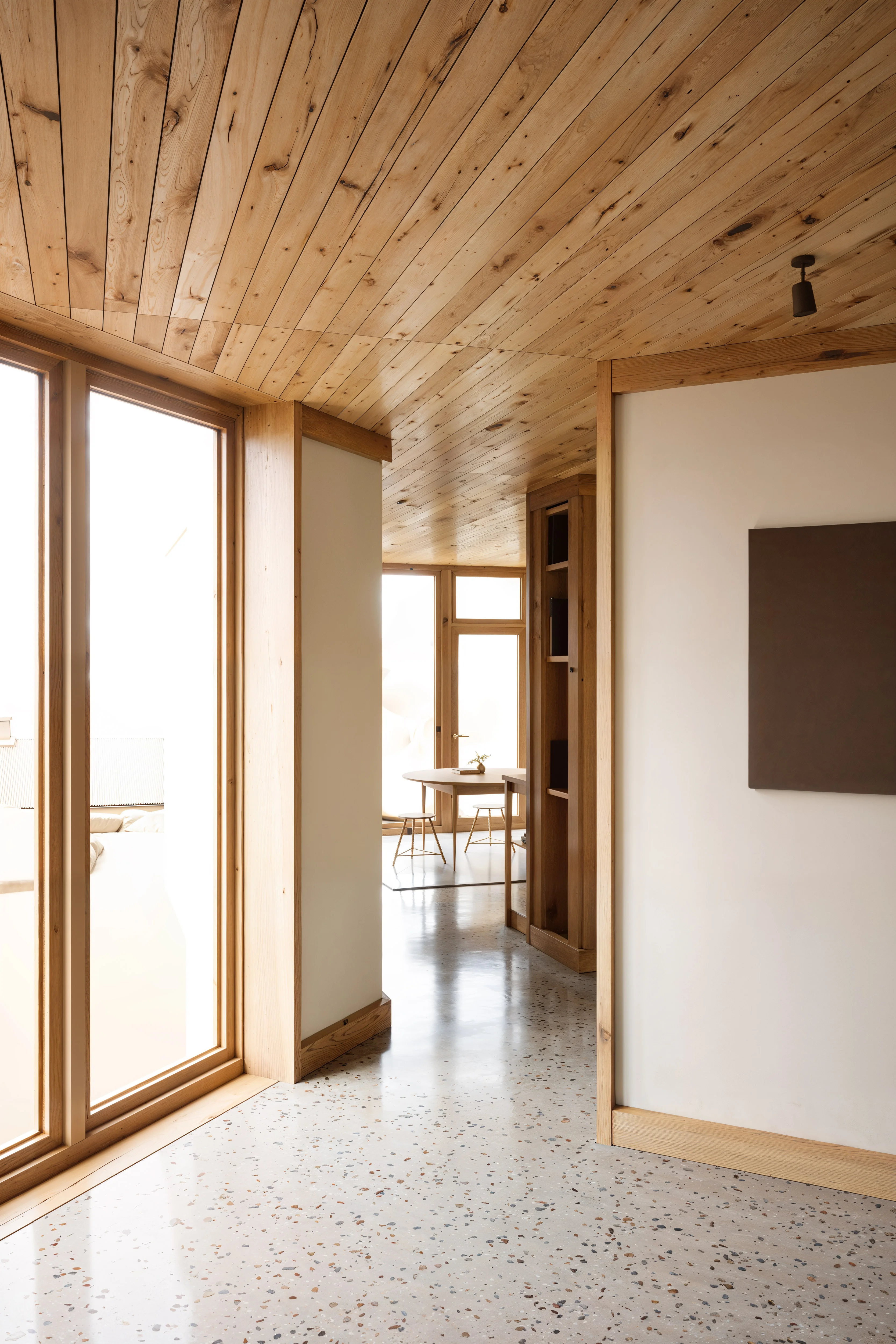 Modern interior hallway with wooden ceiling, large windows, terrazzo floor, and a dining table with stools in the background.