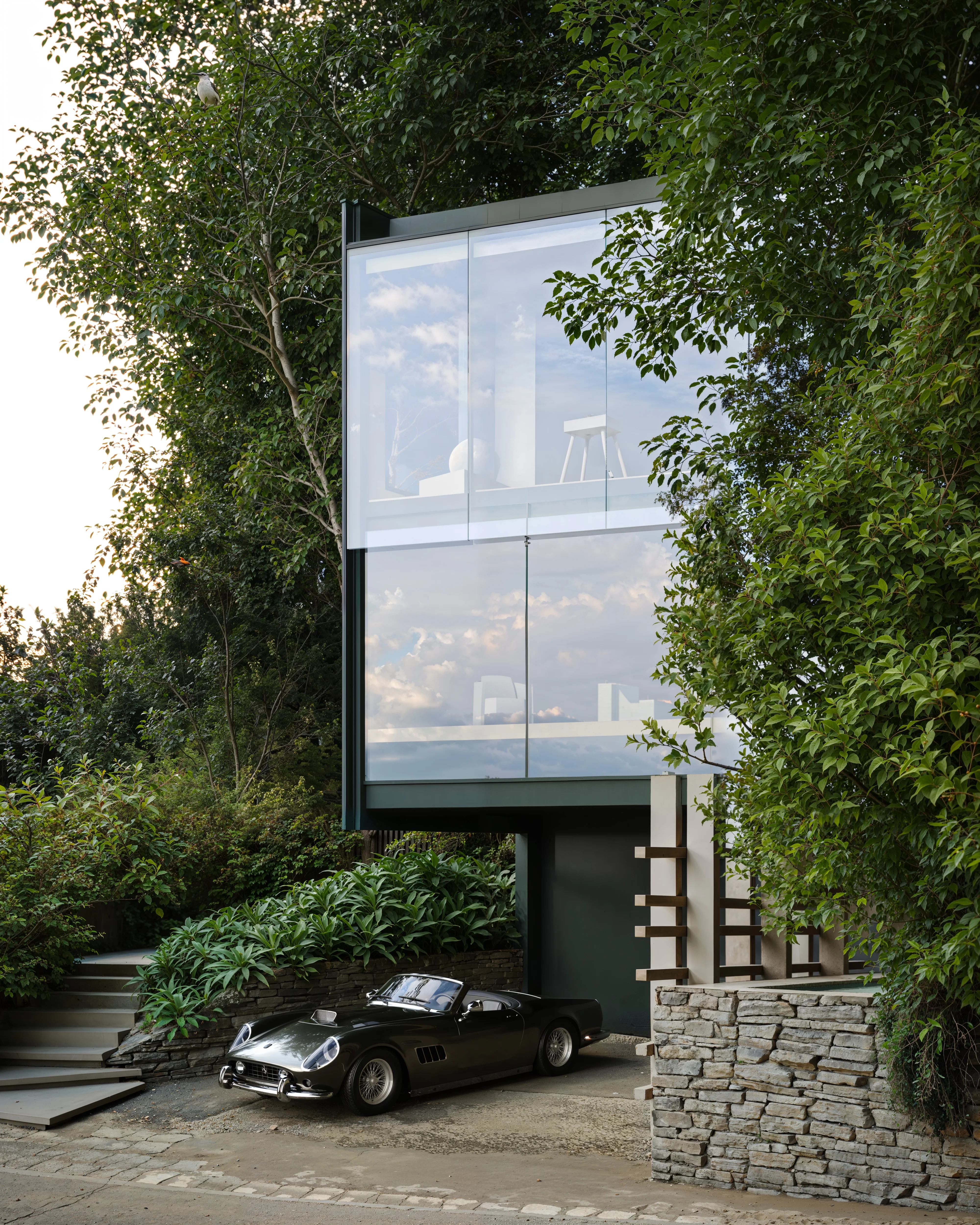 Modern house with large glass windows surrounded by dense greenery and a vintage black convertible car parked in front.