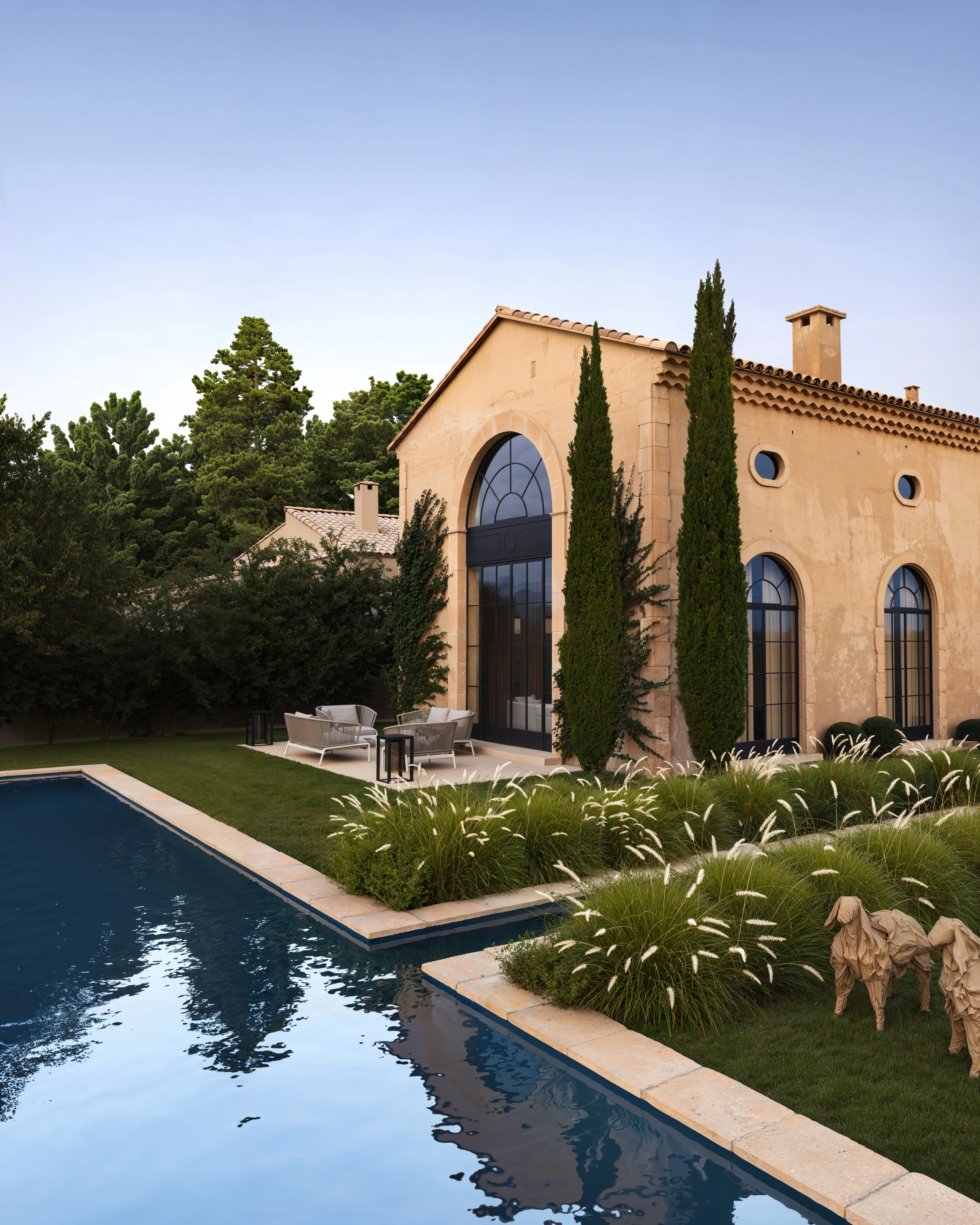 Mediterranean-style house with arched windows, tall cypress trees, a patio with outdoor furniture, and a swimming pool reflecting the clear sky.