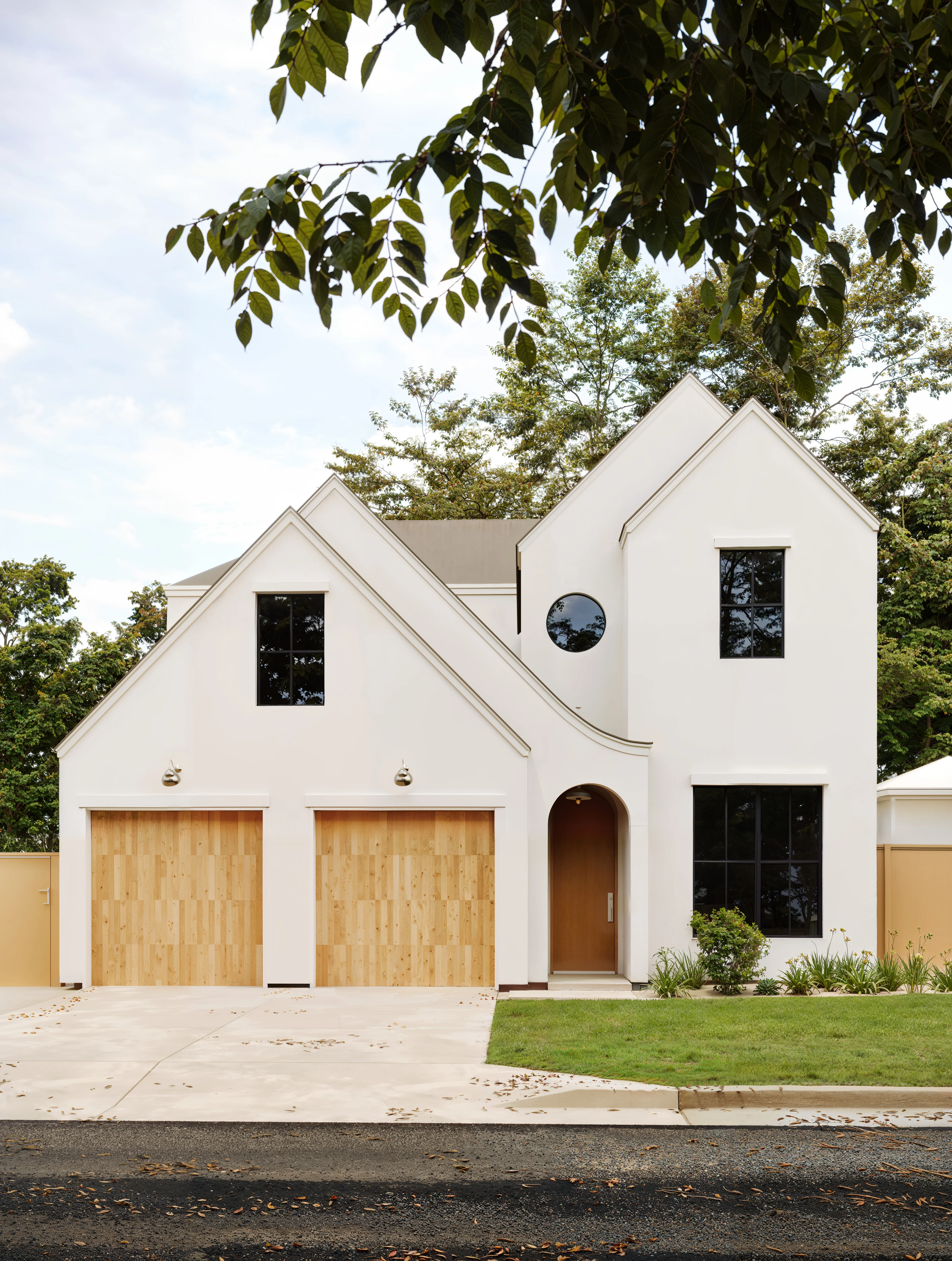 Modern white two-story house with two wooden garage doors, black-framed windows, and an arched front door under tree leaves.