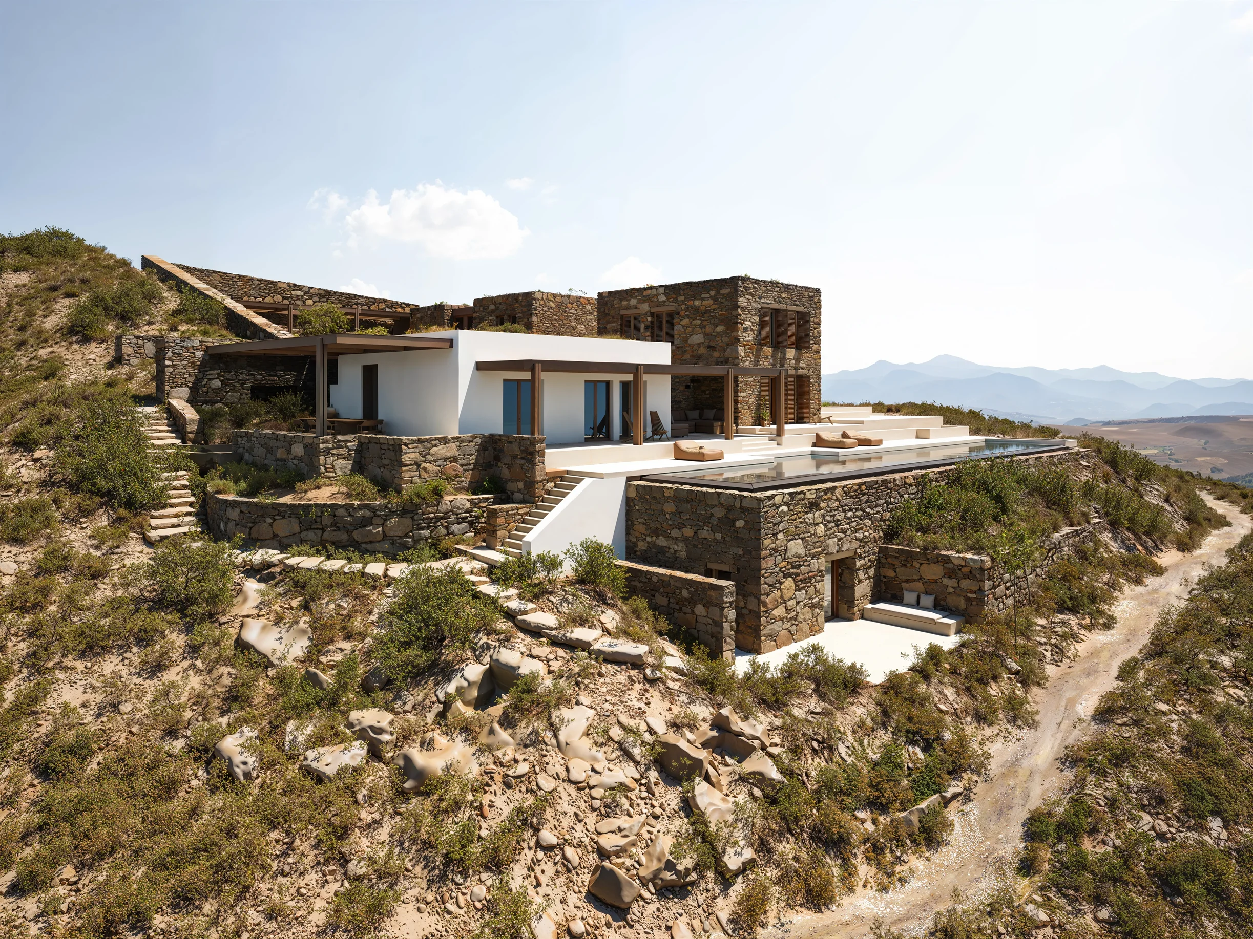 Modern hillside villa with stone and white walls, large windows, terraces, and an infinity pool overlooking a mountainous landscape.