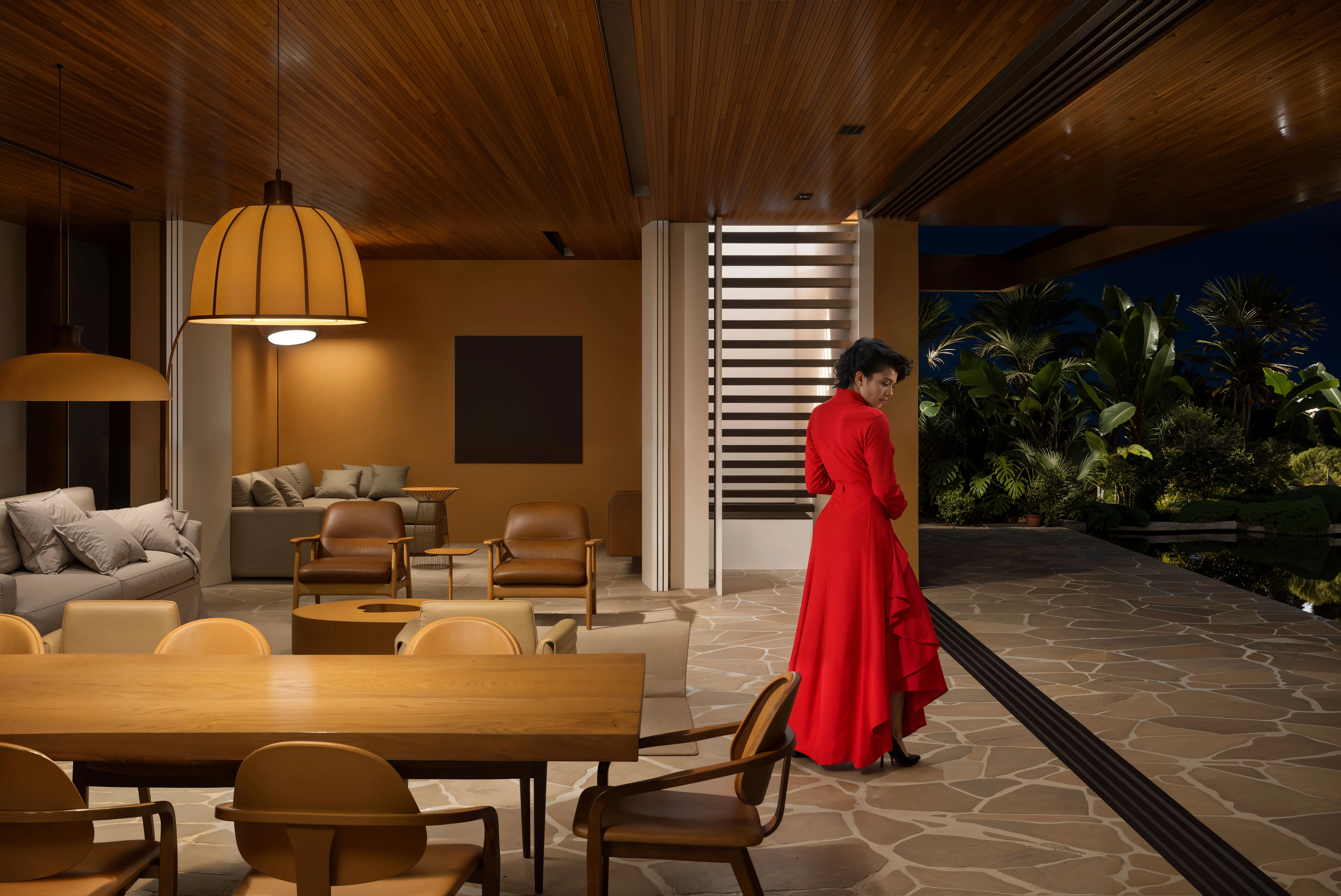 Woman in a long red dress standing in a modern indoor-outdoor living space at night with brown and beige furniture and lush greenery outside.