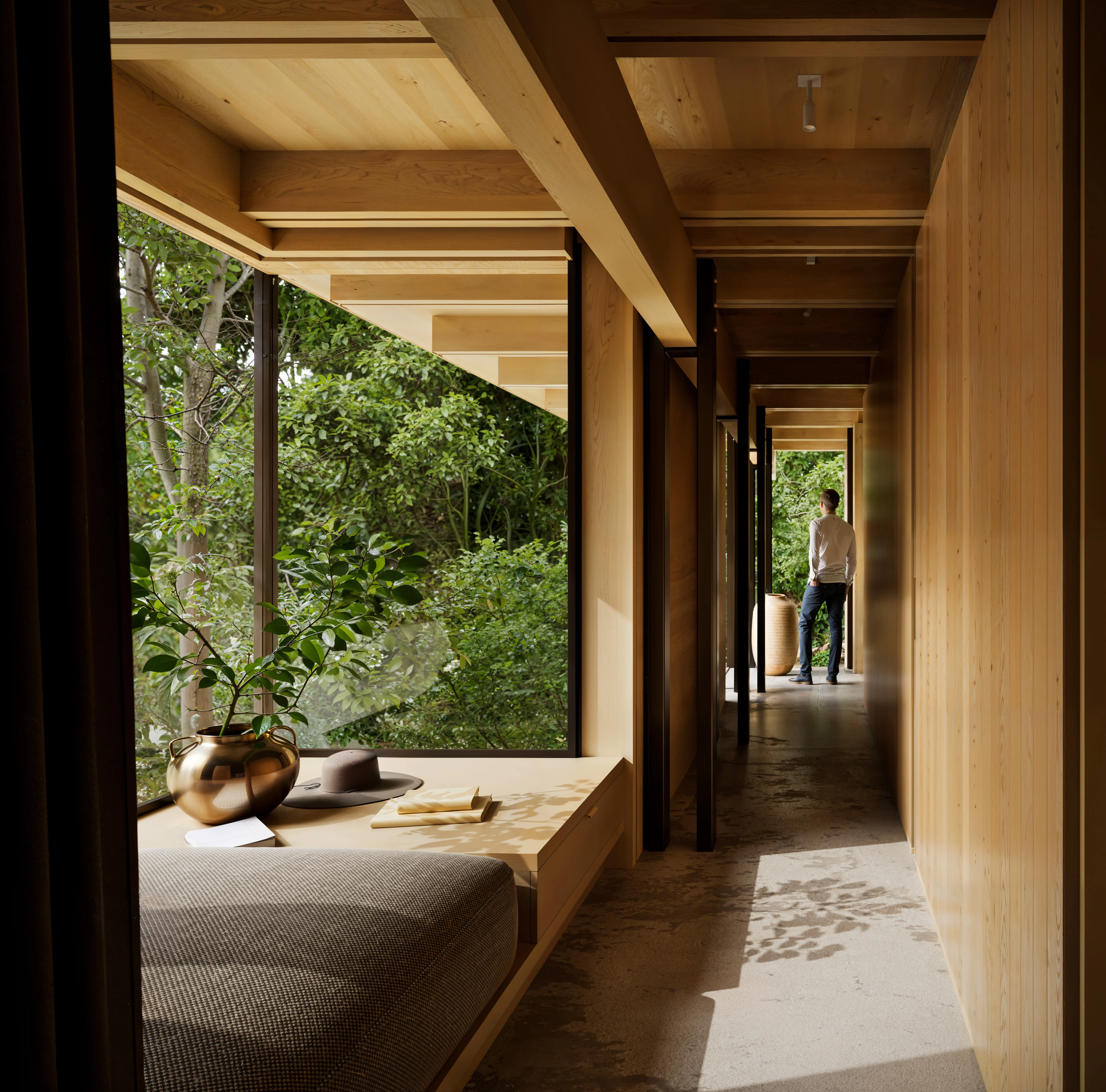 Sunlit wooden hallway with large windows overlooking green trees, a cushioned bench with a brass vase, hat, and books, and a person standing at the end looking outside.
