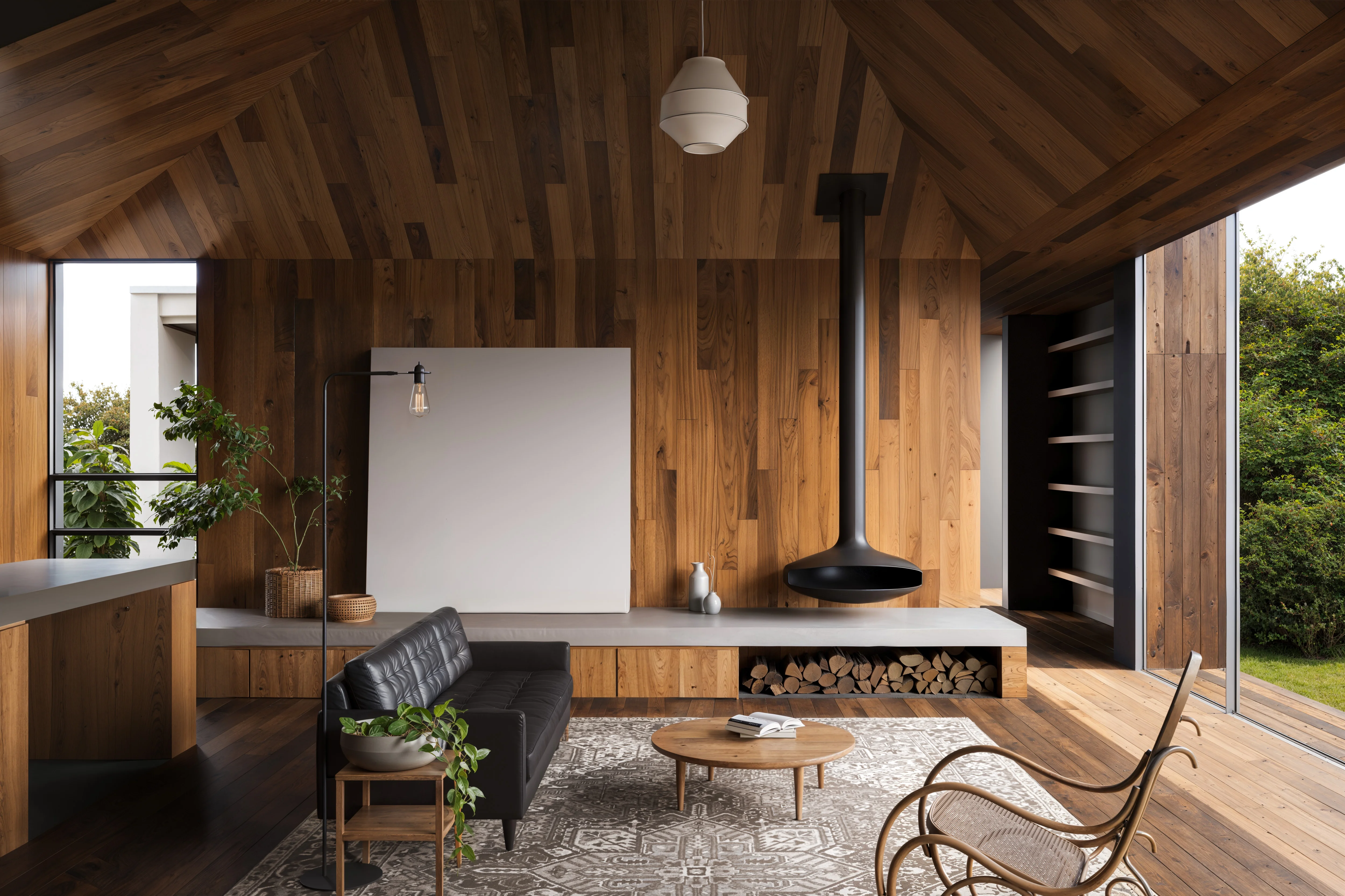 Modern living room with wood-paneled walls and ceiling, a black leather sofa, suspended black fireplace, round wooden coffee table, patterned rug, and large windows with garden view.
