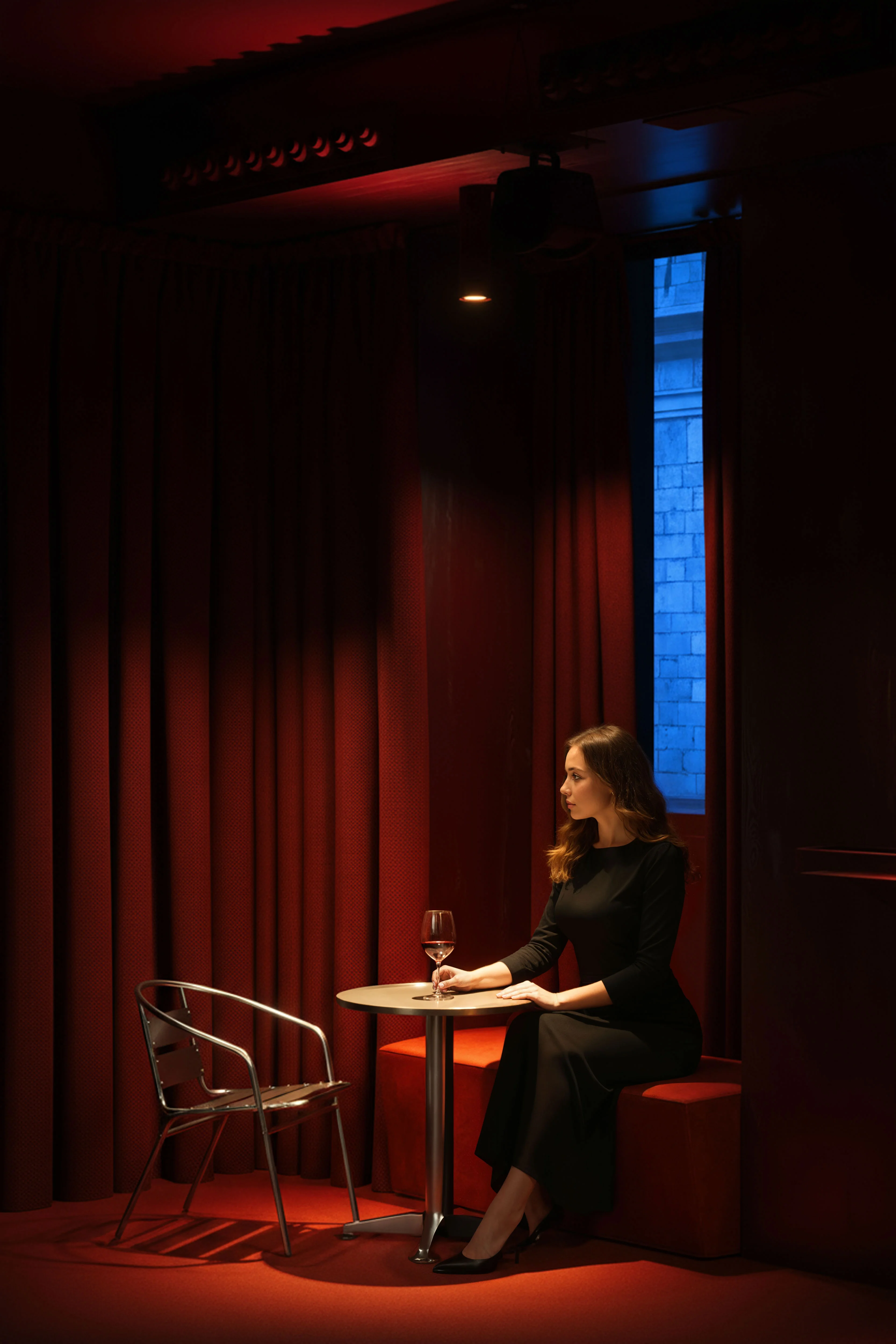 Woman in a black dress sitting alone at a small round table in a dimly lit red room holding a wine glass.