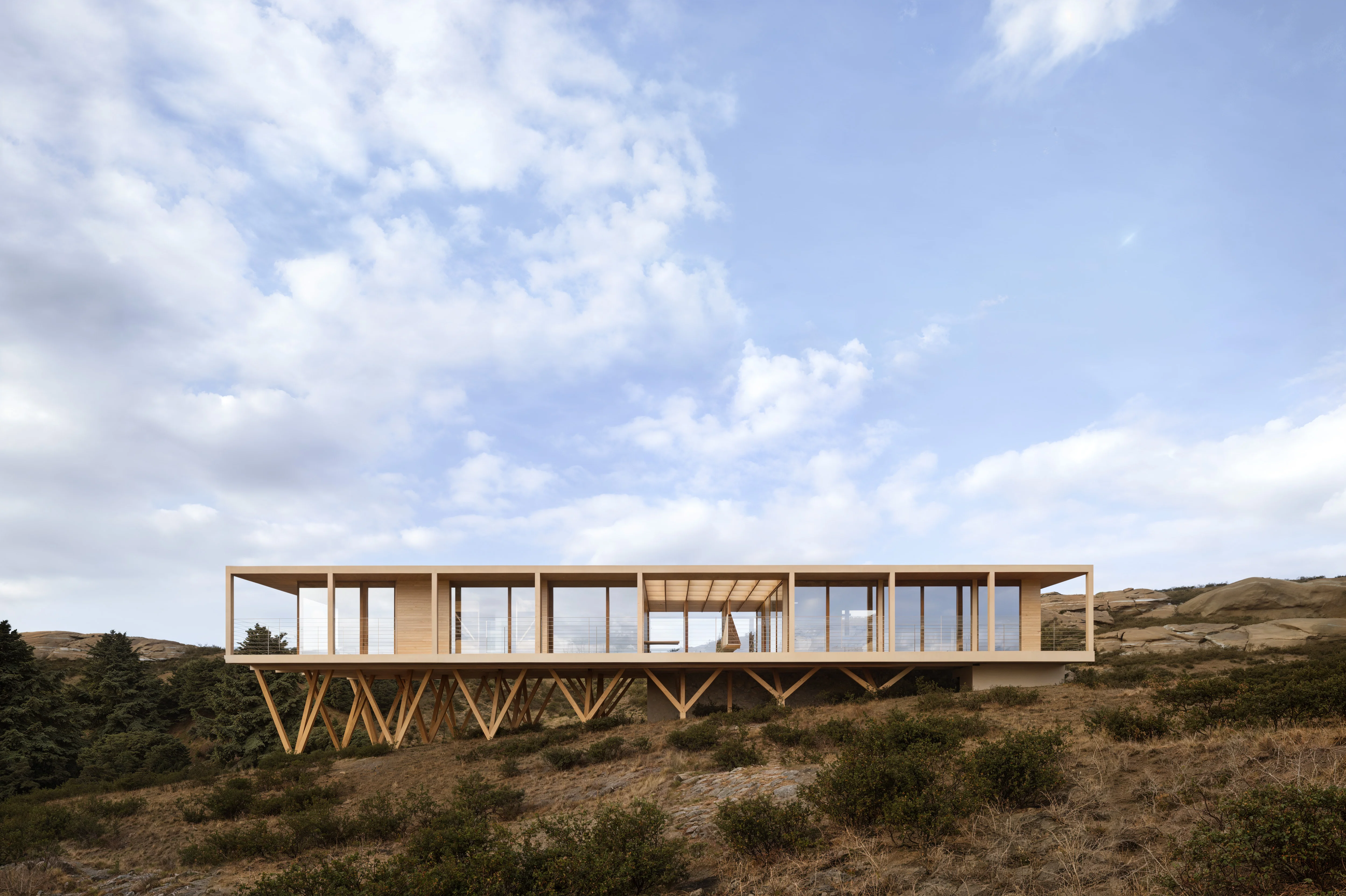 Modern elevated wooden house with large glass windows on a sloped, grassy hillside under a partly cloudy sky.