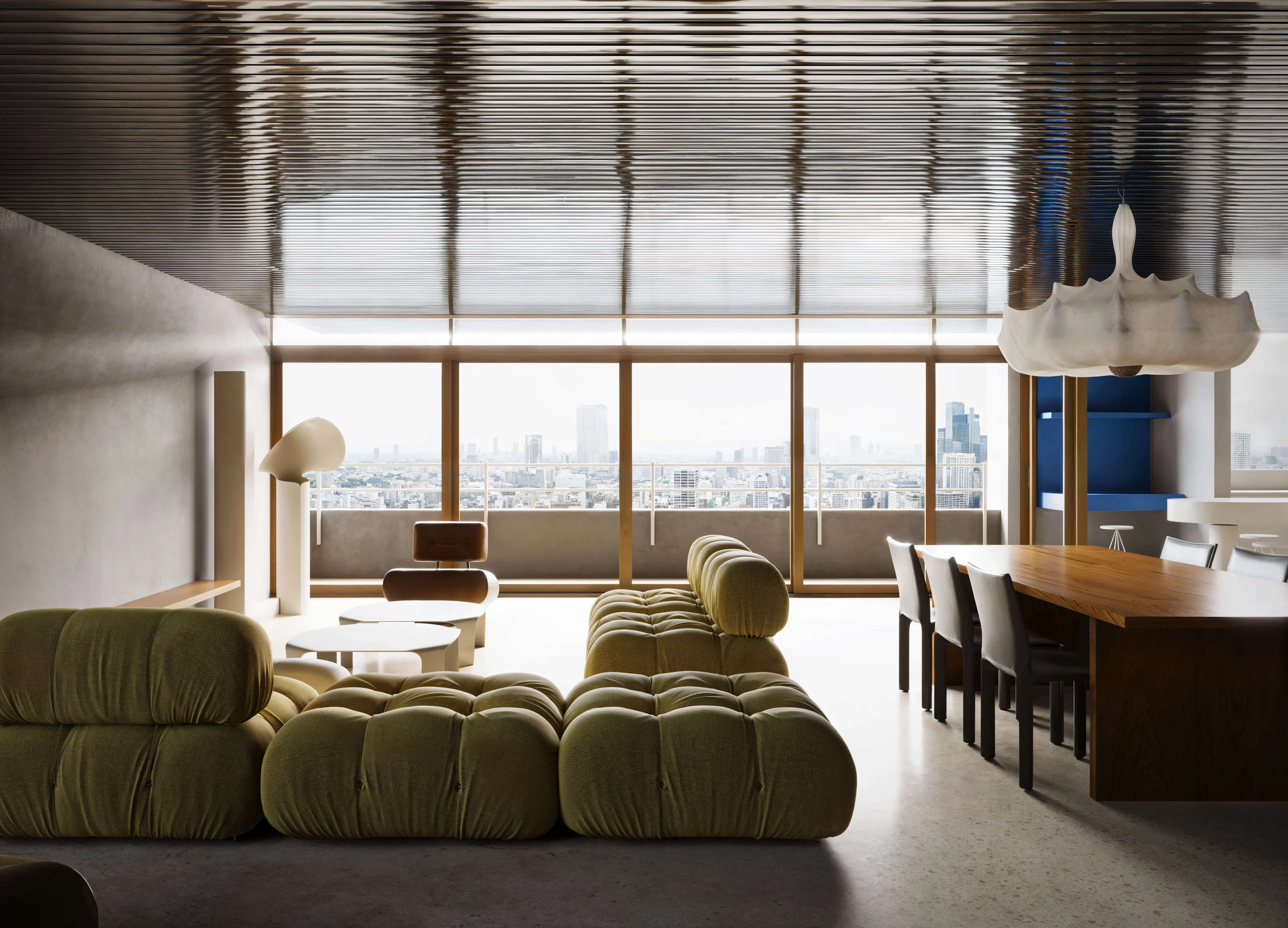 Modern living room with tufted olive-green sectional sofa, wooden dining table with six chairs, large floor lamp, and city skyline view through floor-to-ceiling windows.