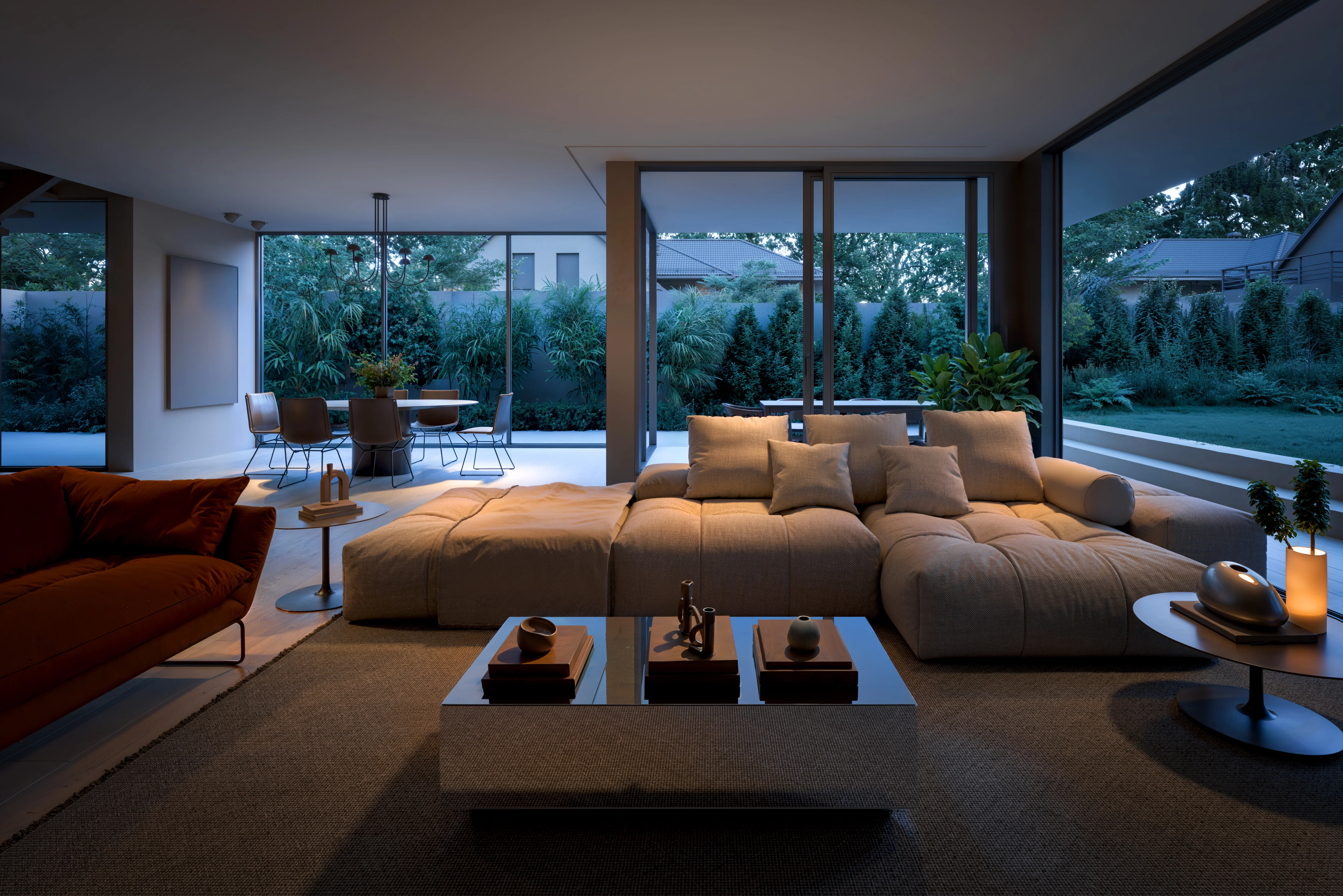 Modern living room with large beige sectional sofa, glass coffee table, and large windows overlooking a garden and outdoor dining area at dusk.