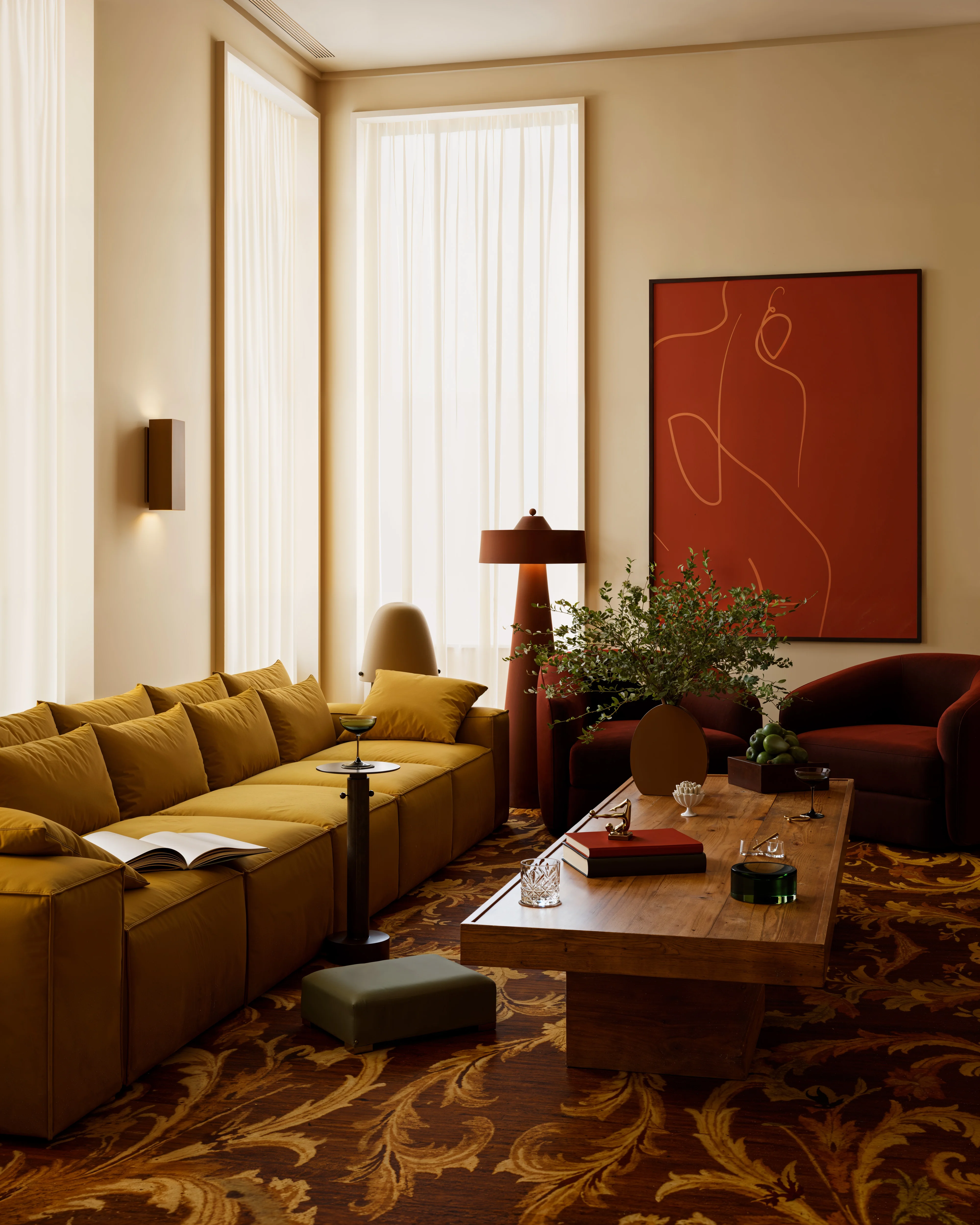Living room with mustard yellow sofa, wooden coffee table with books and fruit, two burgundy armchairs, and large abstract art on beige wall.