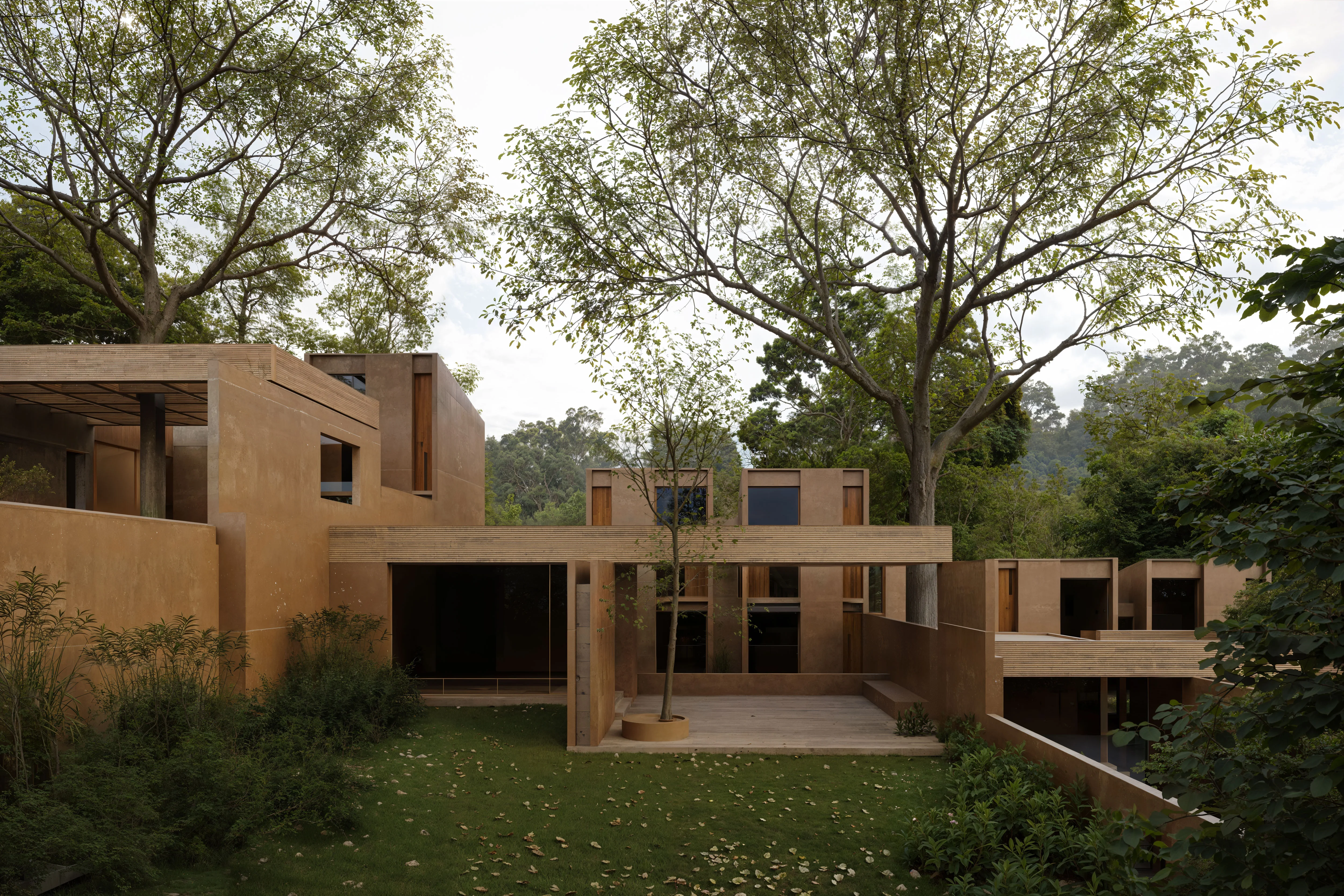 Modern brown concrete building with wooden accents surrounded by green trees and plants under an overcast sky.