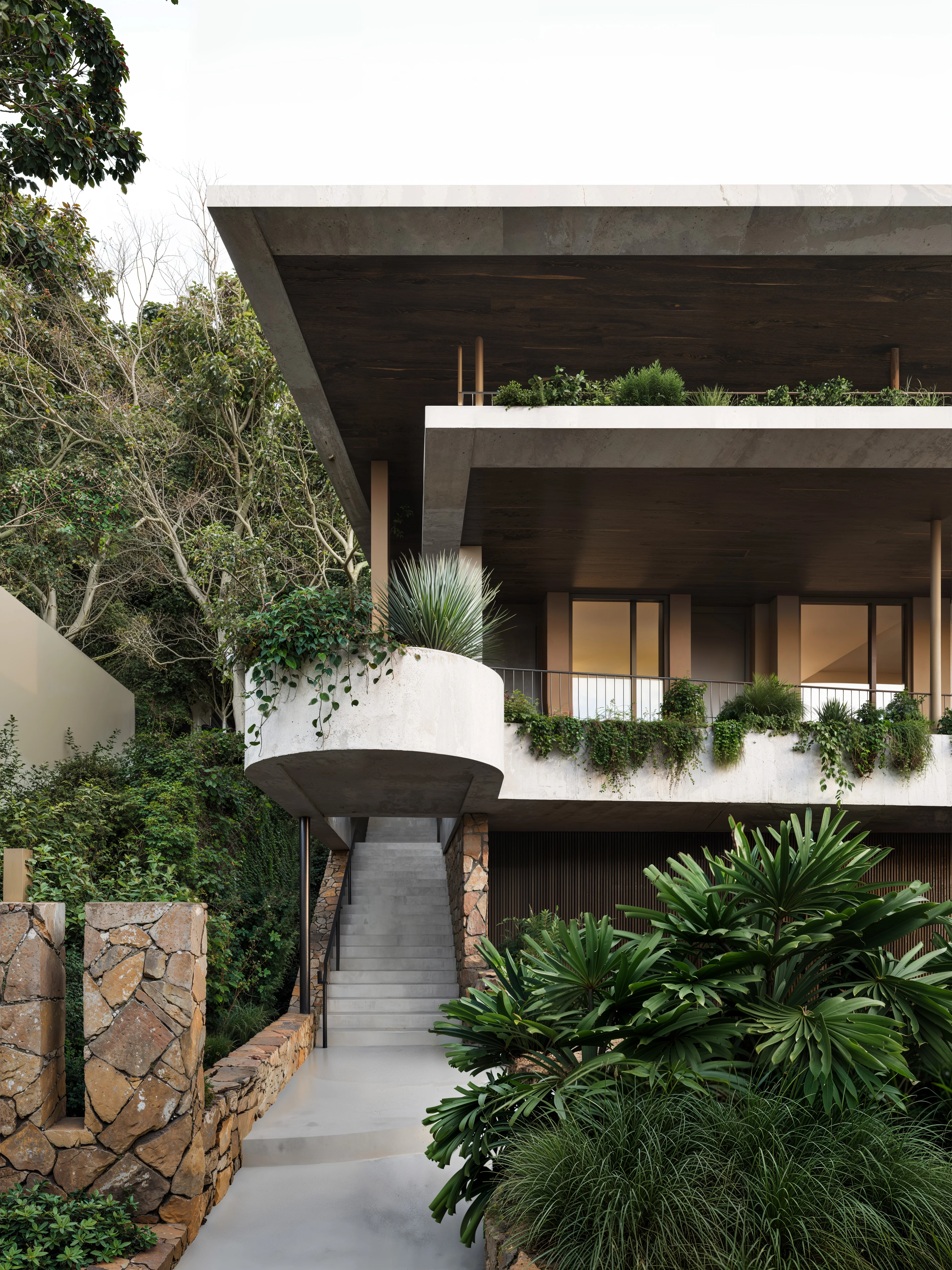 Modern multi-level house with concrete and stone elements, featuring stairway entrance and lush greenery around balconies and garden.