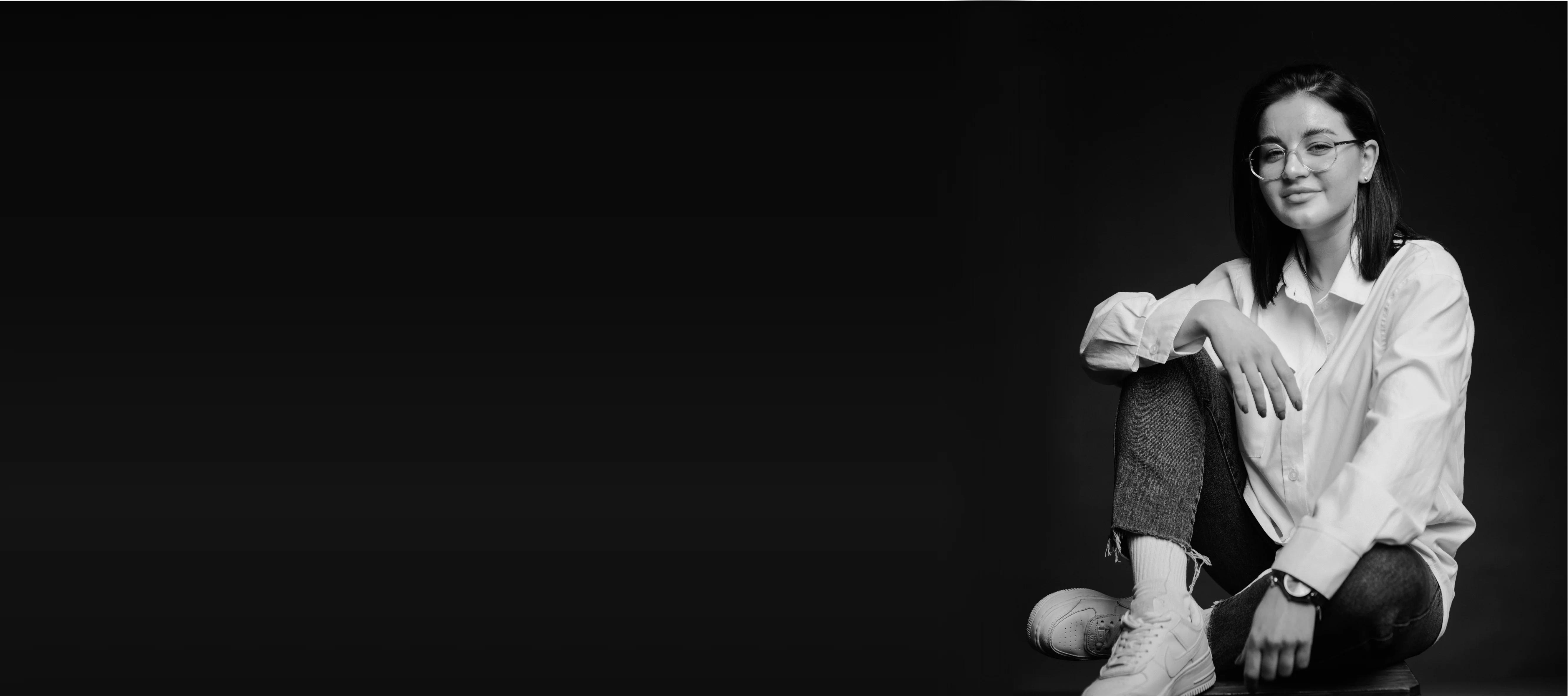 Young woman with glasses sitting cross-legged on a stool against a dark background, wearing a white shirt, jeans, and white sneakers.