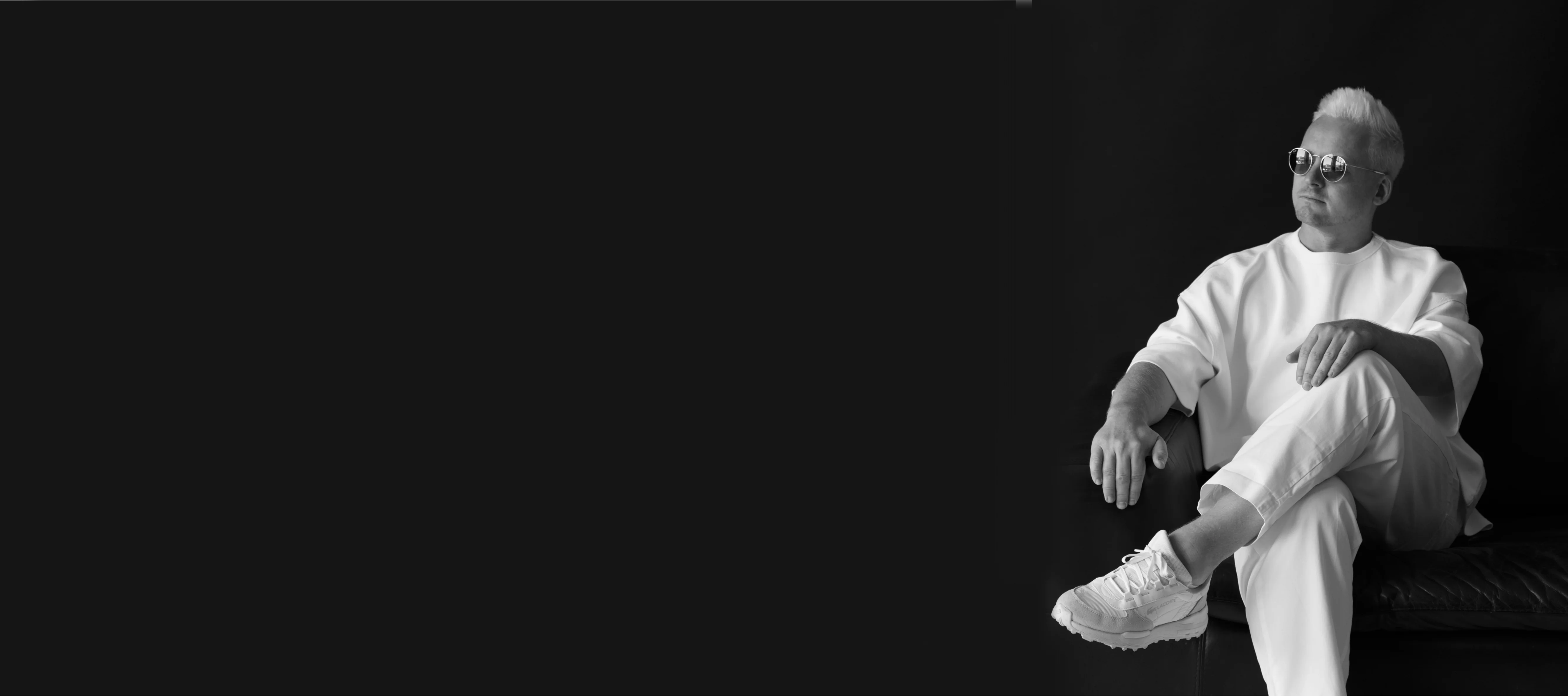 Man with short light hair wearing sunglasses, white sweatshirt, pants, and sneakers sitting with crossed legs on a dark couch against a black background.