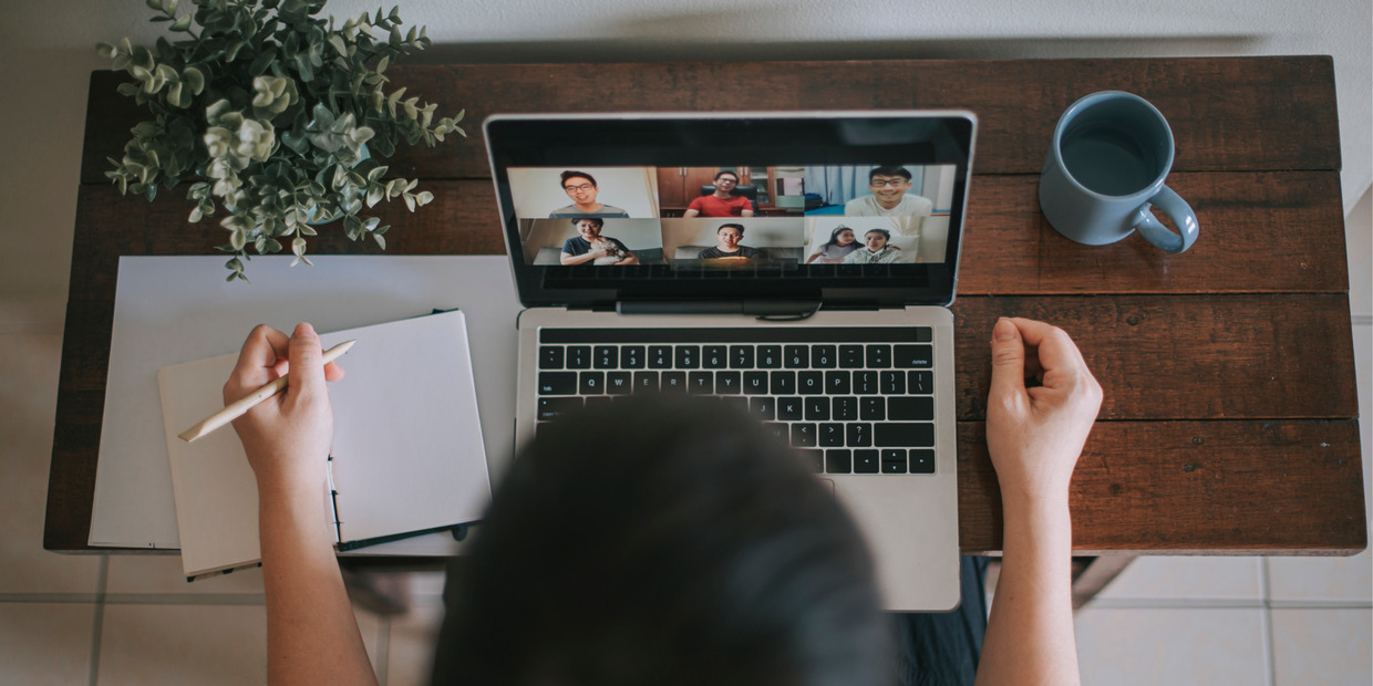 Overhead view of a person taking notes during a video conference on a laptop at a wooden desk with a notebook, coffee mug, and plant nearby.