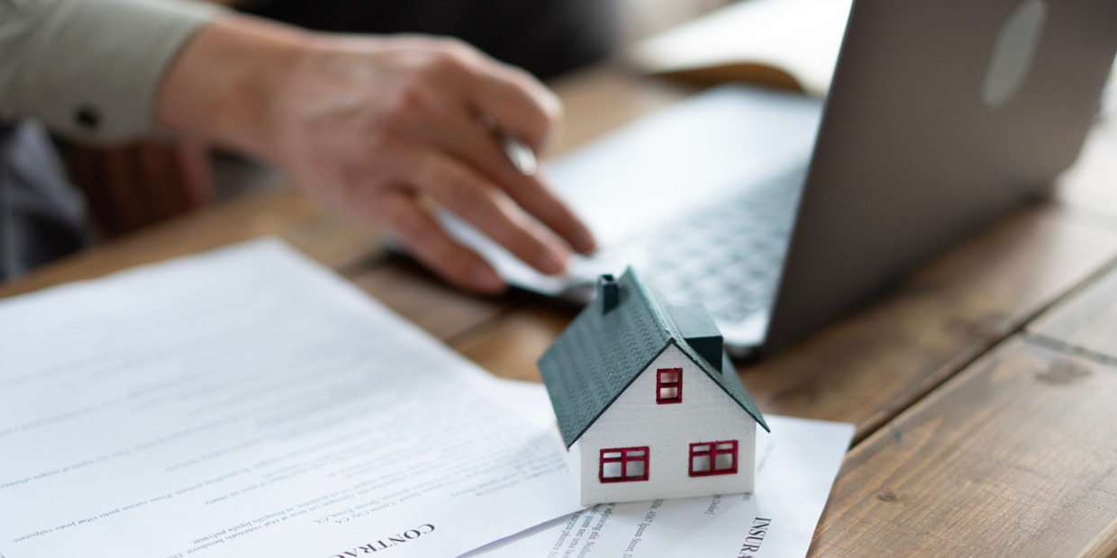 Small model house sitting on insurance documents beside a laptop, with a person working in the background at a wooden desk.