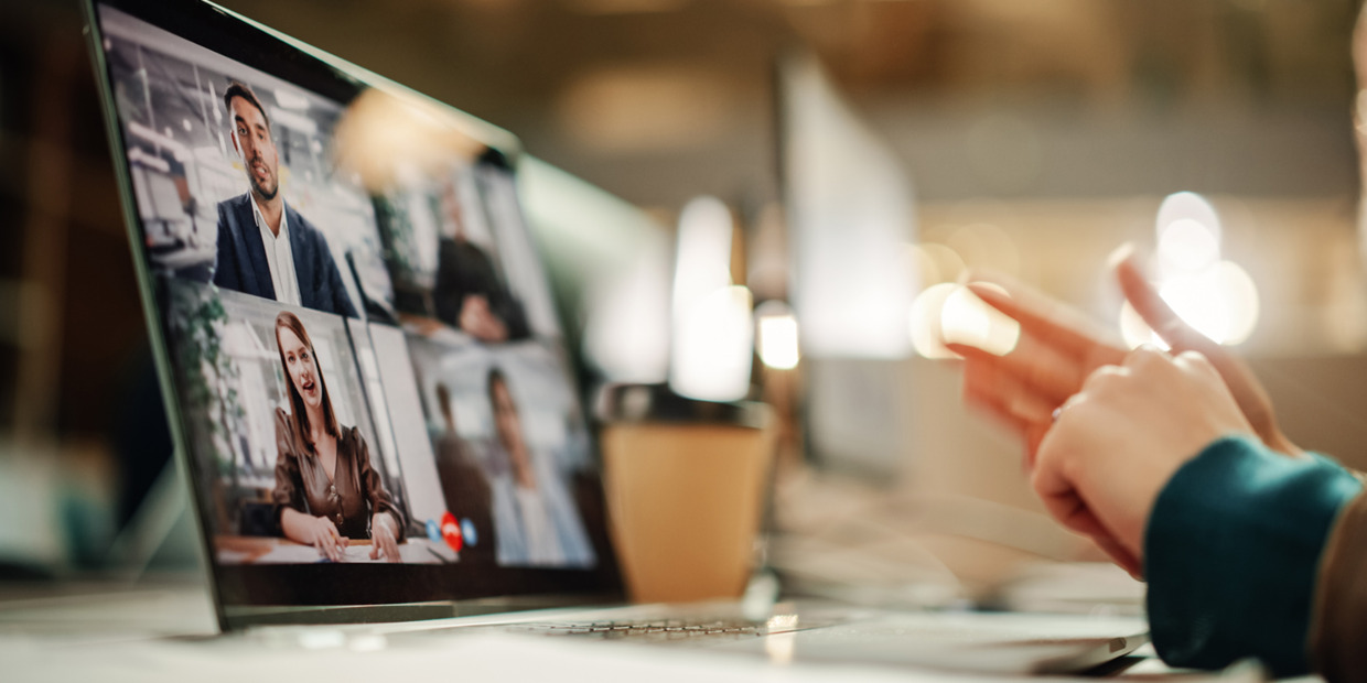 Person participating in a virtual meeting on a laptop, gesturing toward the screen while multiple colleagues appear in a video conference call.