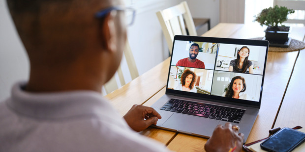 Person participating in a video conference on a laptop at a wooden table, with four attendees visible on screen during a virtual meeting. A smartphone sits nearby, and the bright home setting suggests a remote collaboration or board meeting.