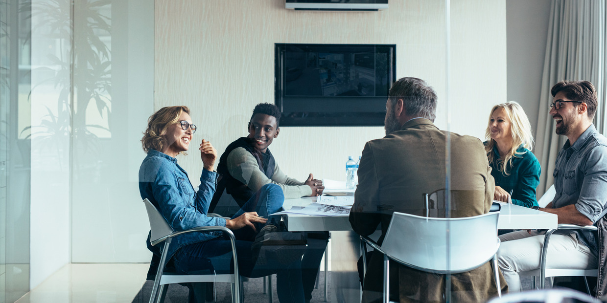 Five colleagues share a discussion and laugh together during a board meeting around a conference table.